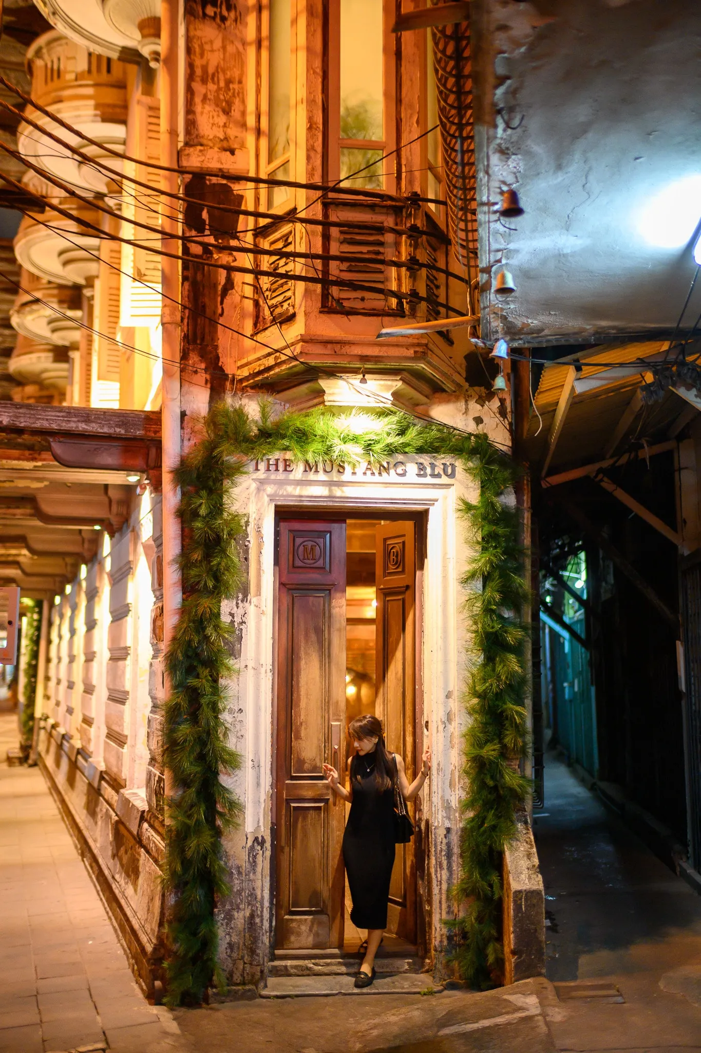 A woman in a black dress standing in an old wooden doorway framed with greenery at night
