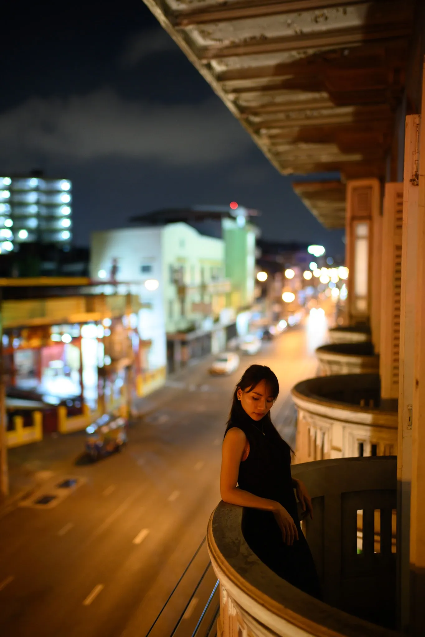 A woman in black leaning on a curved balcony above a lit street at night
