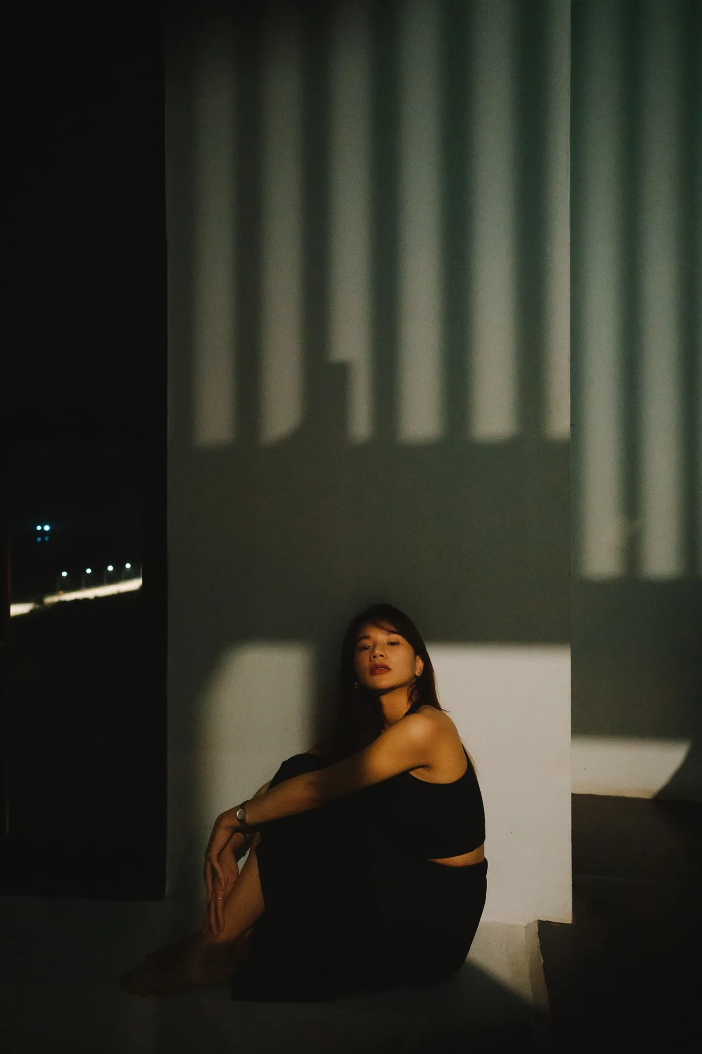 A woman in black sitting in a band of light against a wall striped with shadows