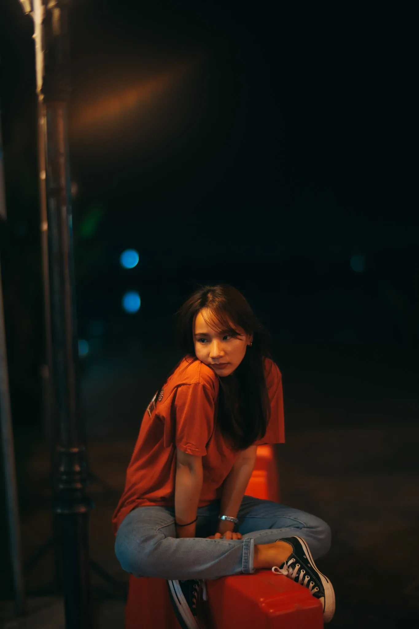 A woman in a red T-shirt and jeans perched on a stool at night