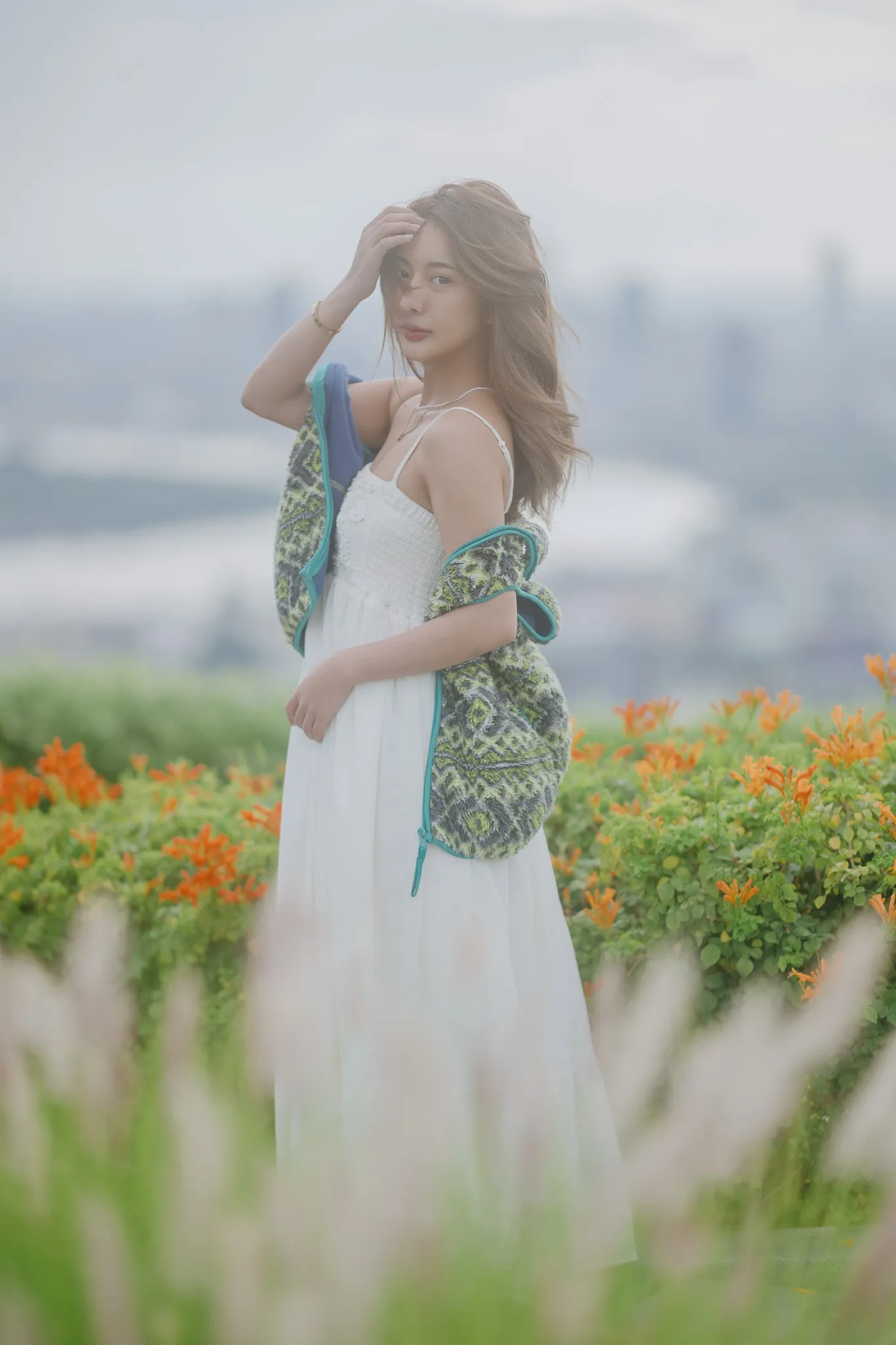 A woman in a white sundress with a patterned shawl standing among orange flowers on a misty hilltop
