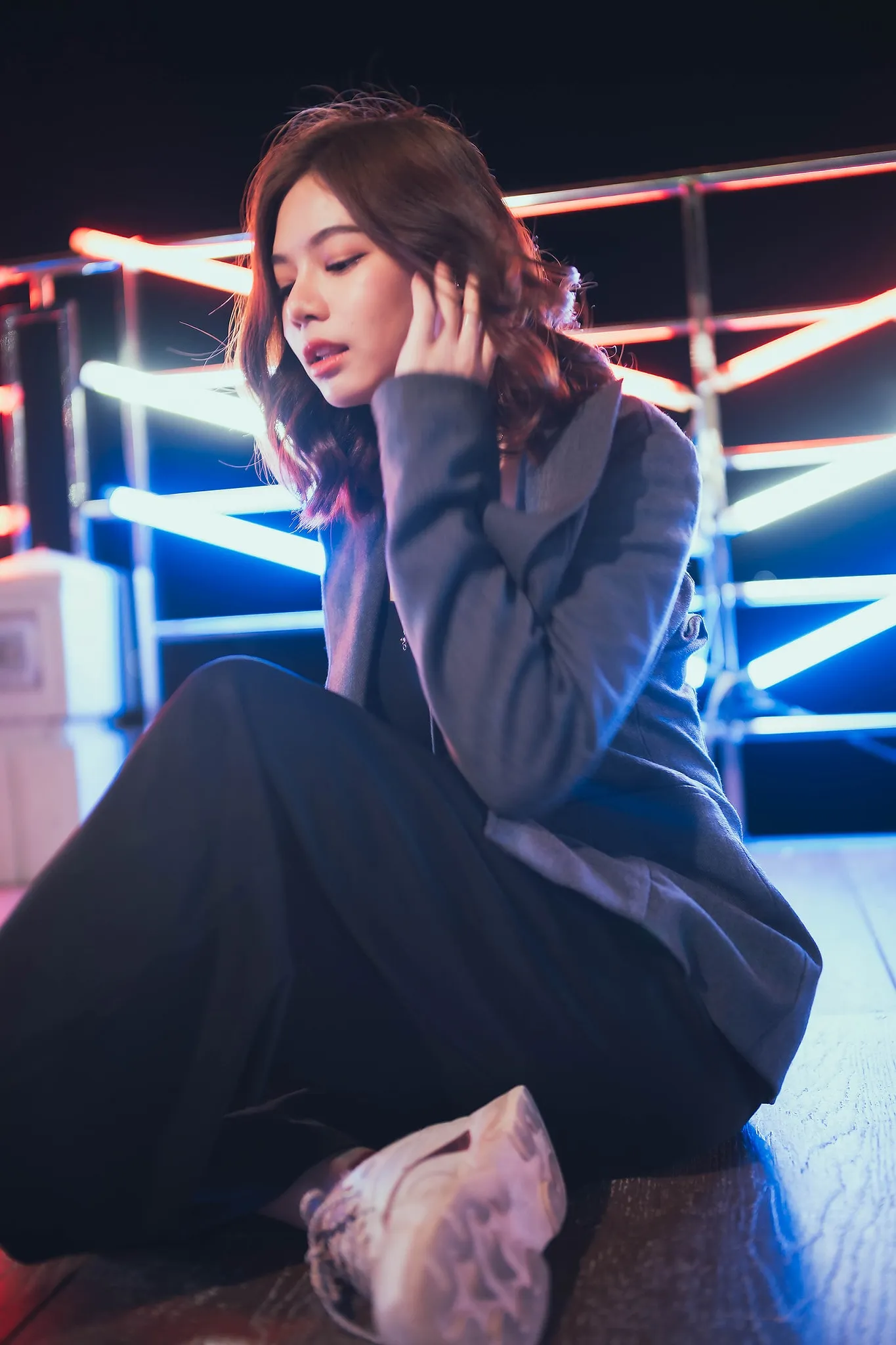 A woman in a grey blazer sitting beside a neon-lit railing at night, hand to her hair