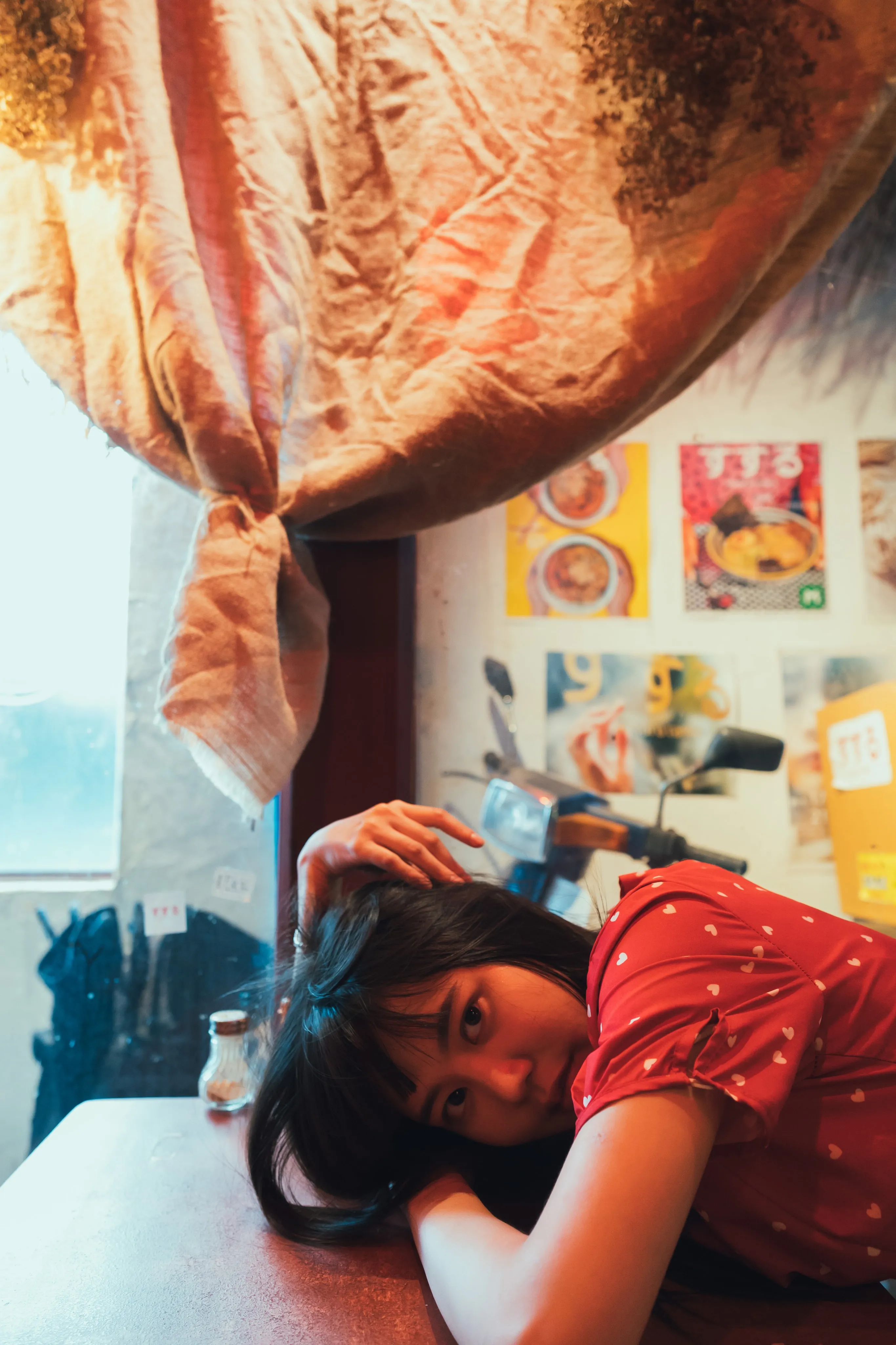 A woman in a red polka-dot dress resting her head on a table in a small café