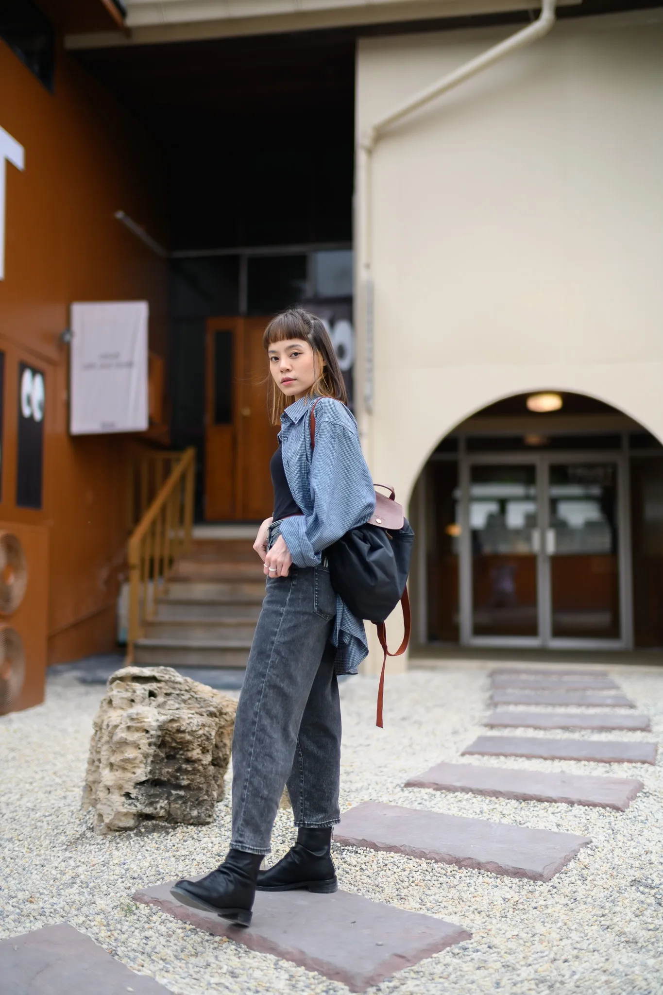 A woman in a denim jacket and backpack standing on stepping stones outside a building