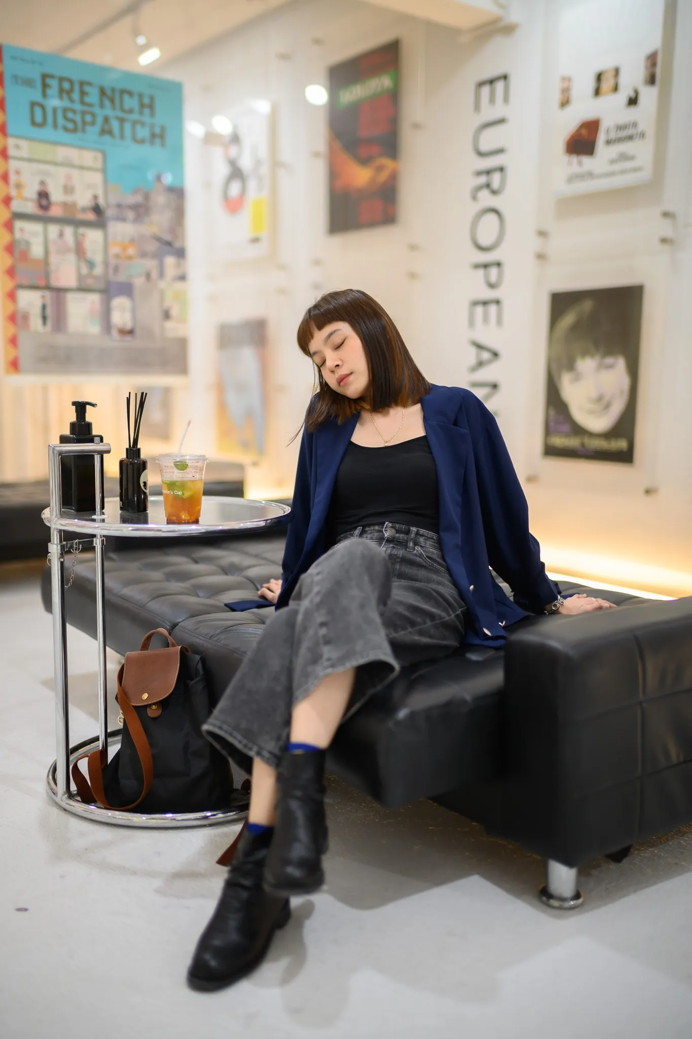 A woman in a navy jacket leaning back on a black leather bench beneath film posters