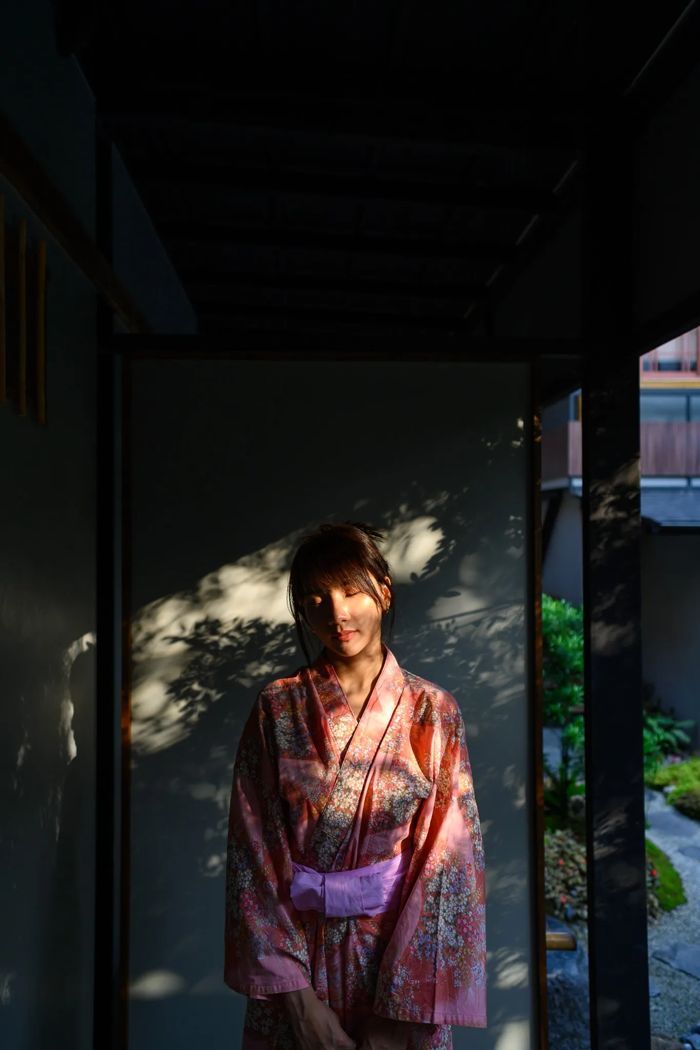 A woman in a pink floral yukata standing in dappled light under a dark wooden eave