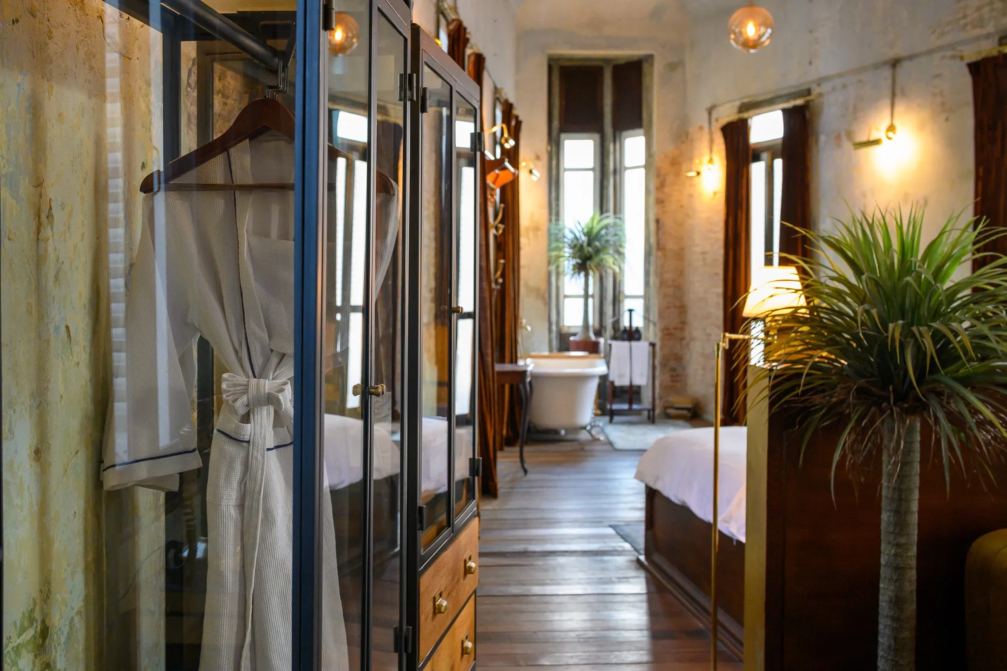 A white robe hanging in a glass wardrobe, with the bedroom and a clawfoot tub beyond