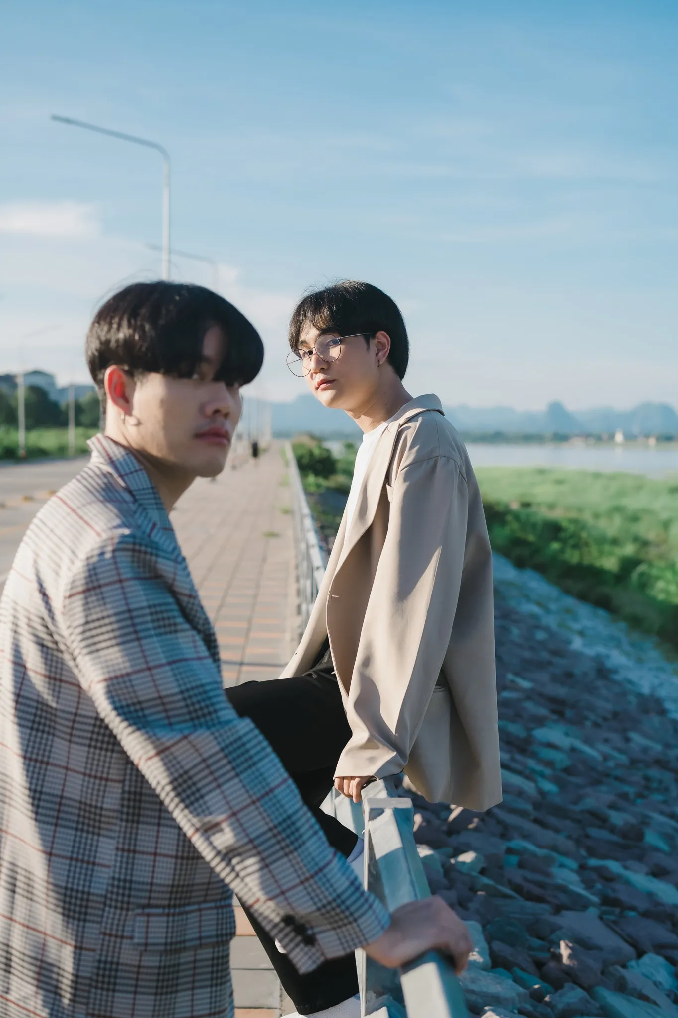 Two young men in checked and beige jackets leaning on a railing along a riverside path