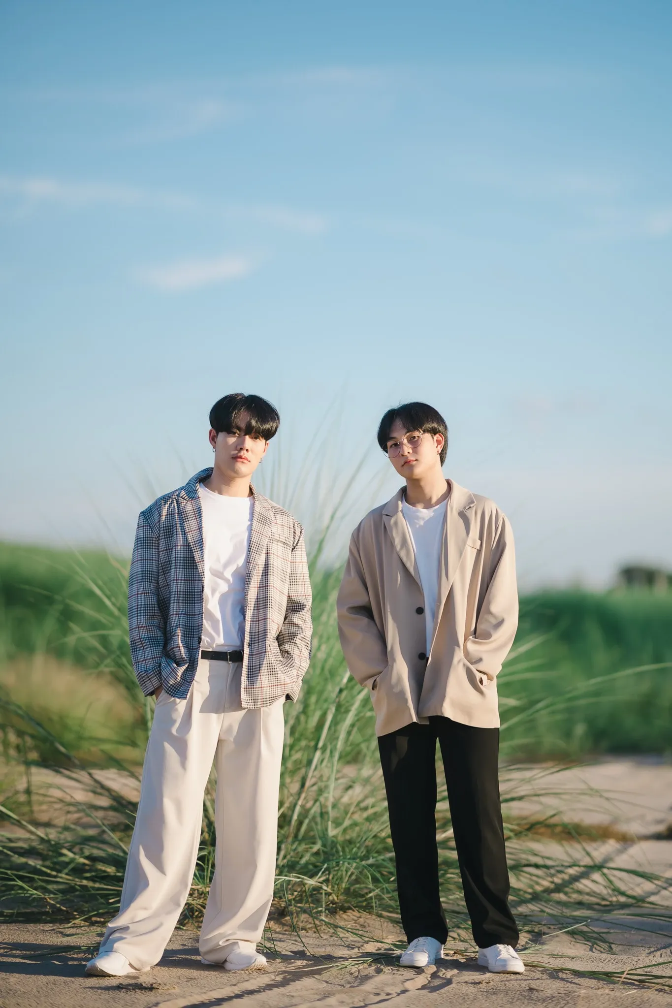 Two young men in checked and beige blazers standing among tall grass under a blue sky