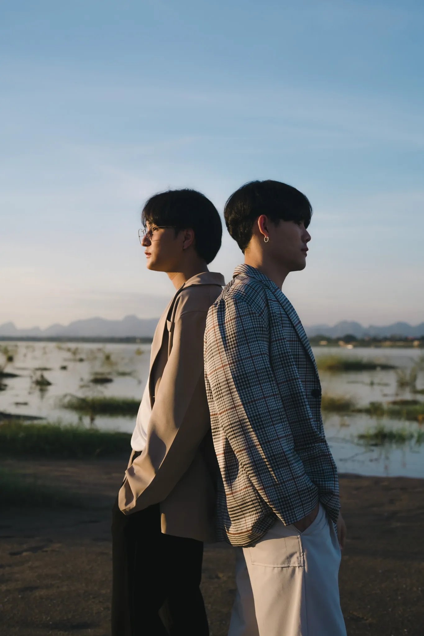 Two young men in beige and checked jackets standing back to back beside a marsh at sunset
