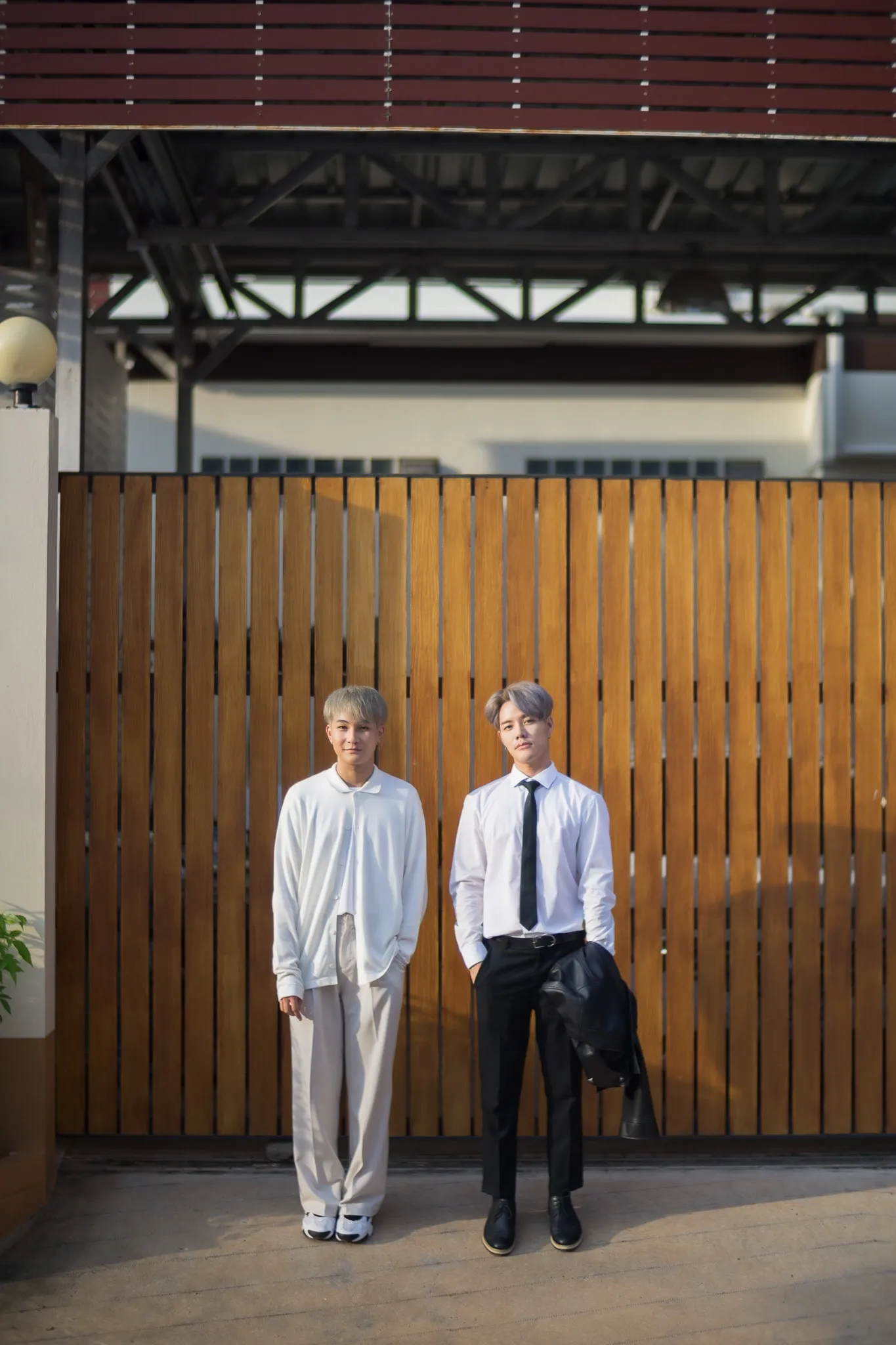 Two young men in white outfits standing in front of a slatted wooden gate