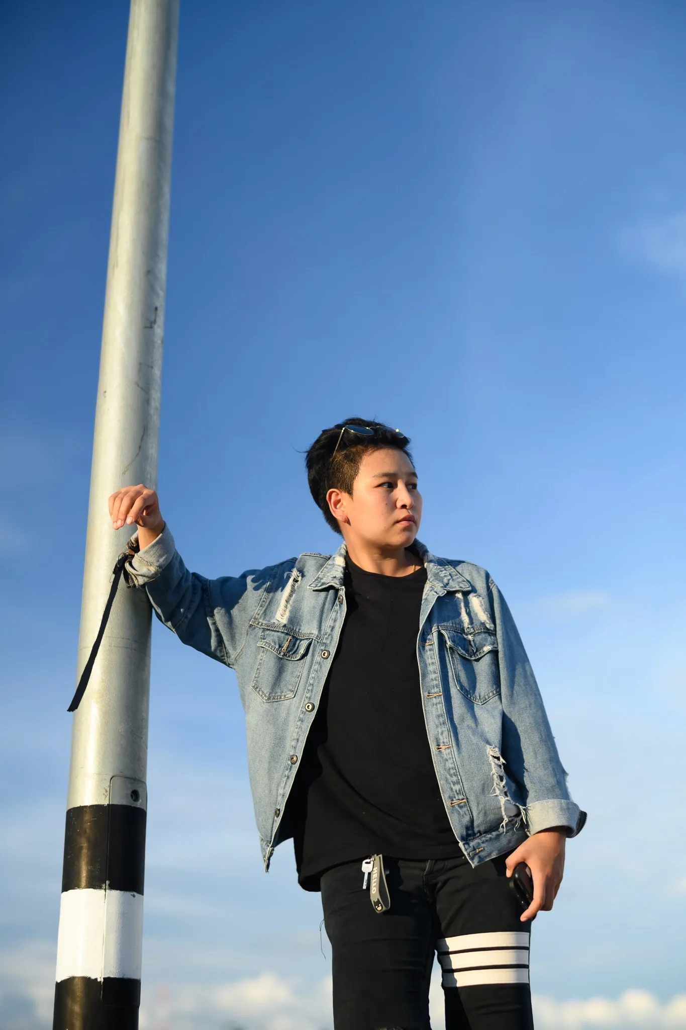 A person in a distressed denim jacket holding a lamppost against a bright blue sky