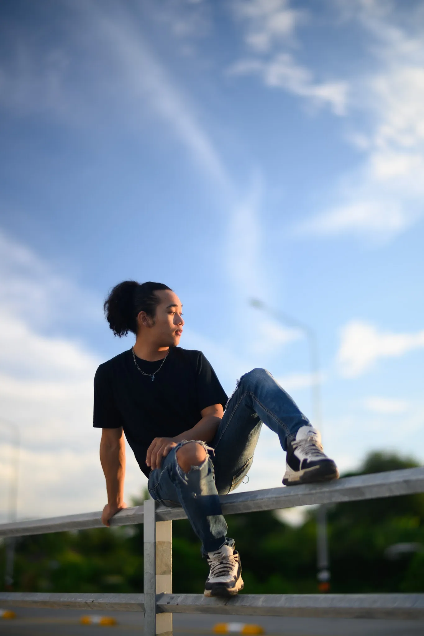 A young man in a black T-shirt and ripped jeans sitting on a metal railing under a blue sky