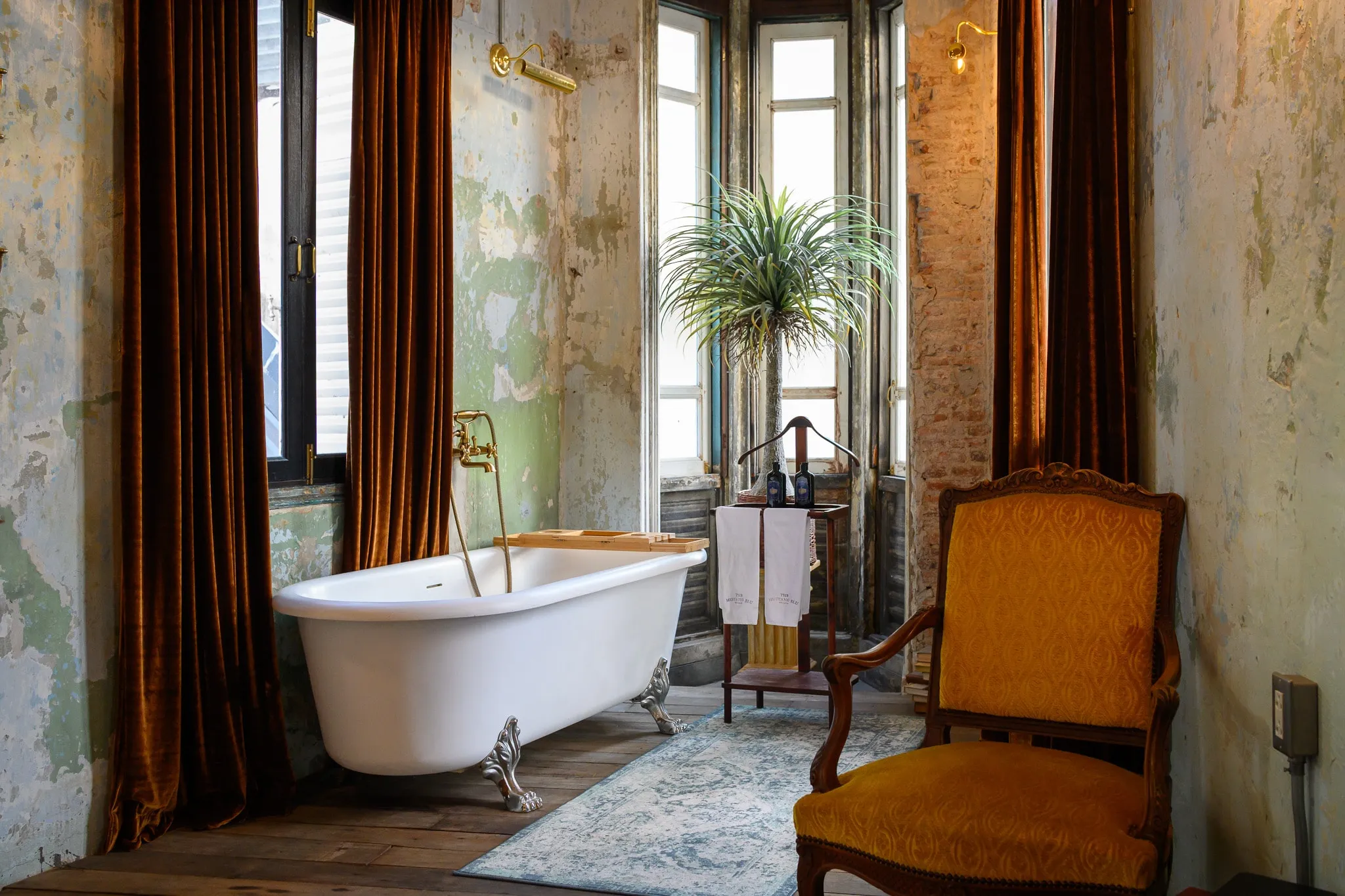 A white clawfoot bathtub beside velvet curtains and a gold armchair in a heritage suite