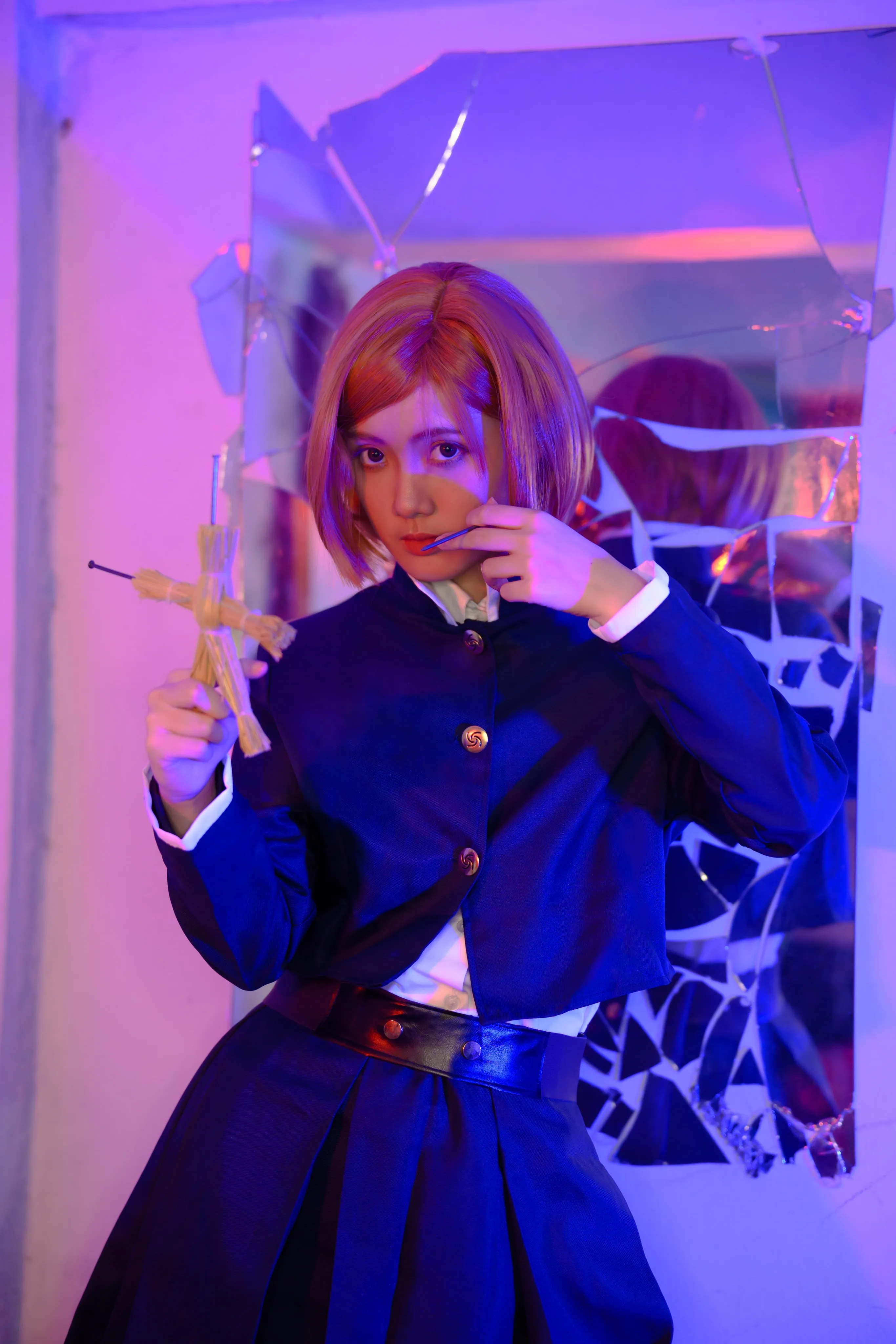 A cosplayer in an orange bob wig and dark school uniform holding a nail and doll before a cracked mirror
