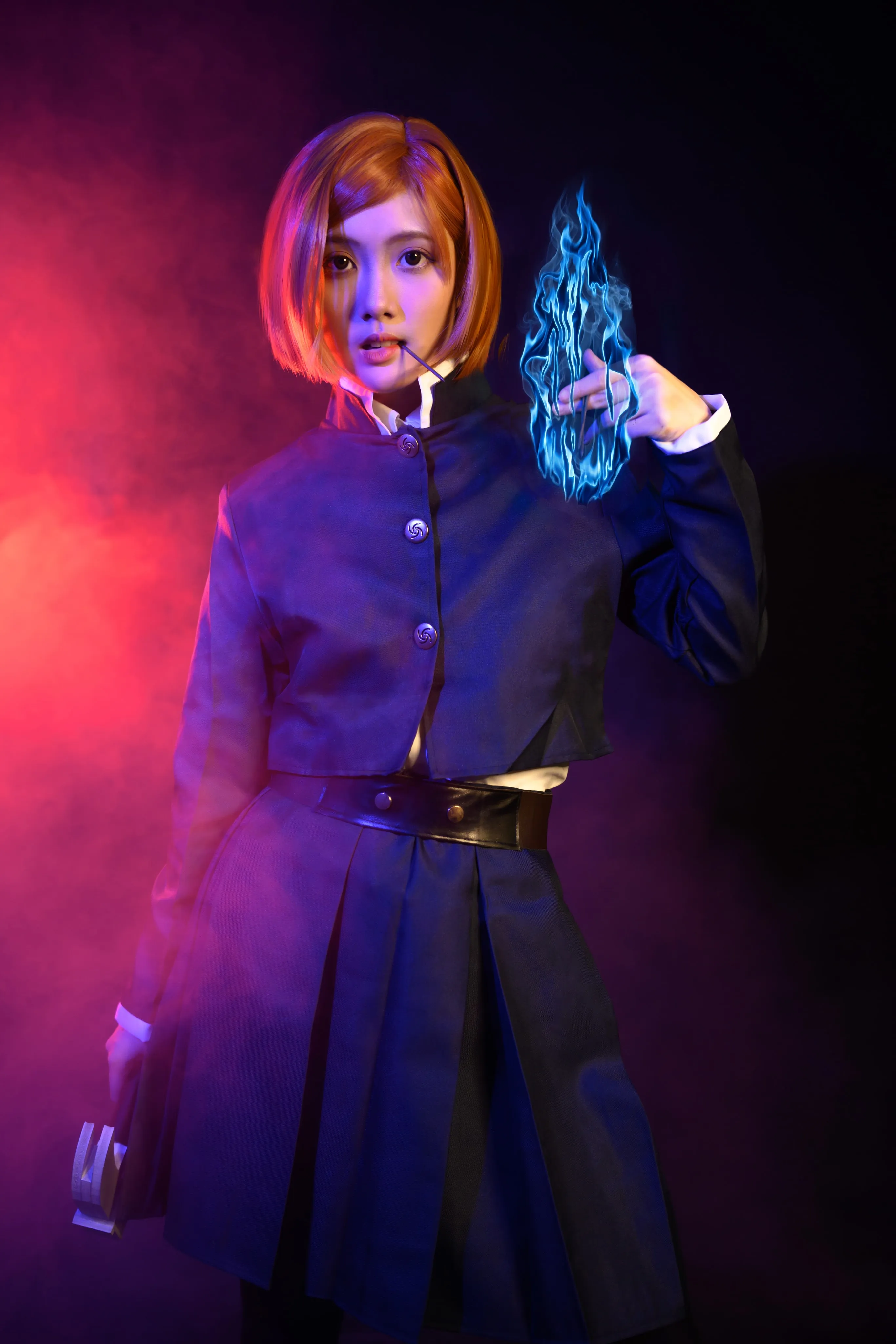 A cosplayer in an orange bob wig and dark uniform holding a blue flame in red smoke