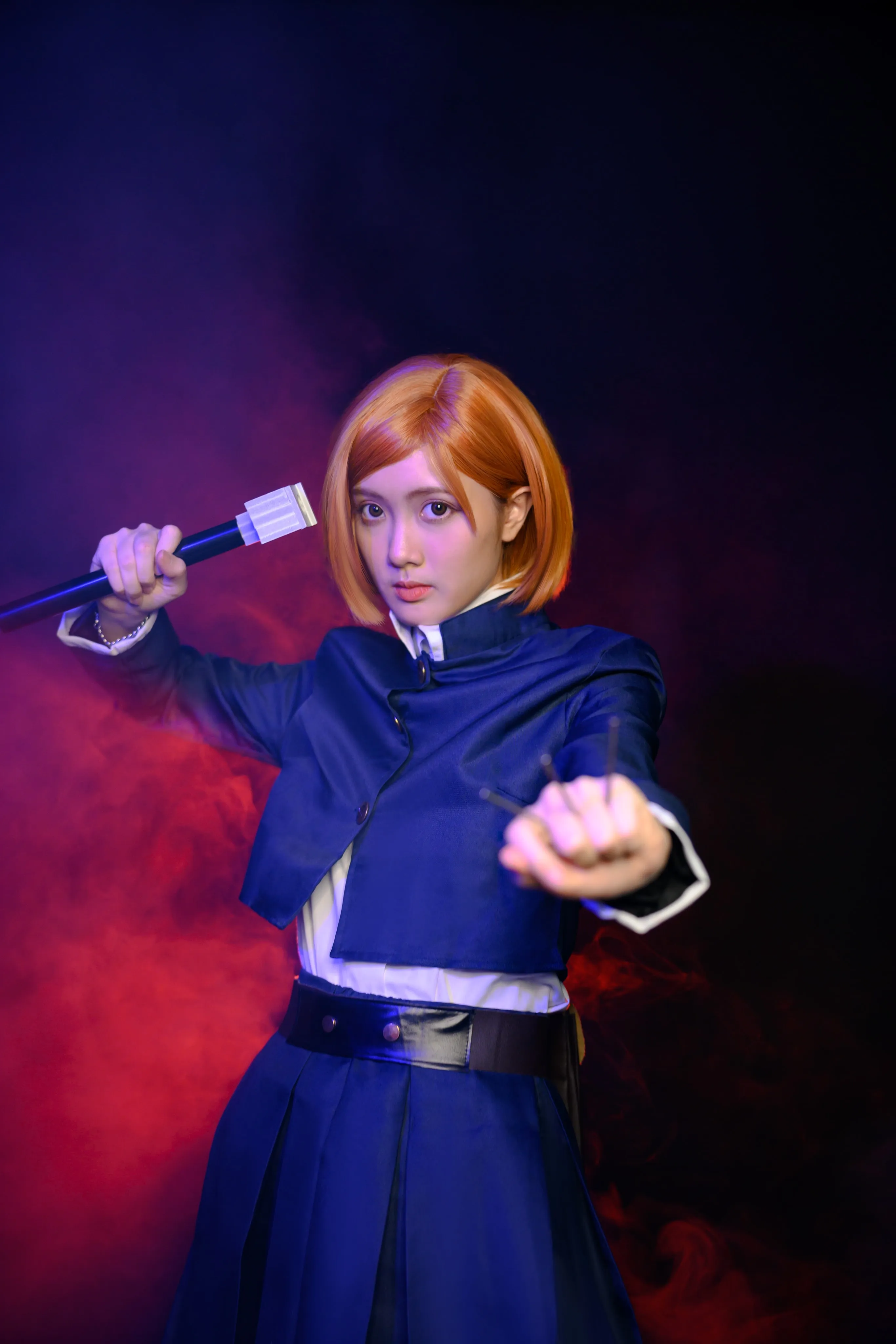 A cosplayer in an orange bob wig and dark uniform raising a hammer against red smoke