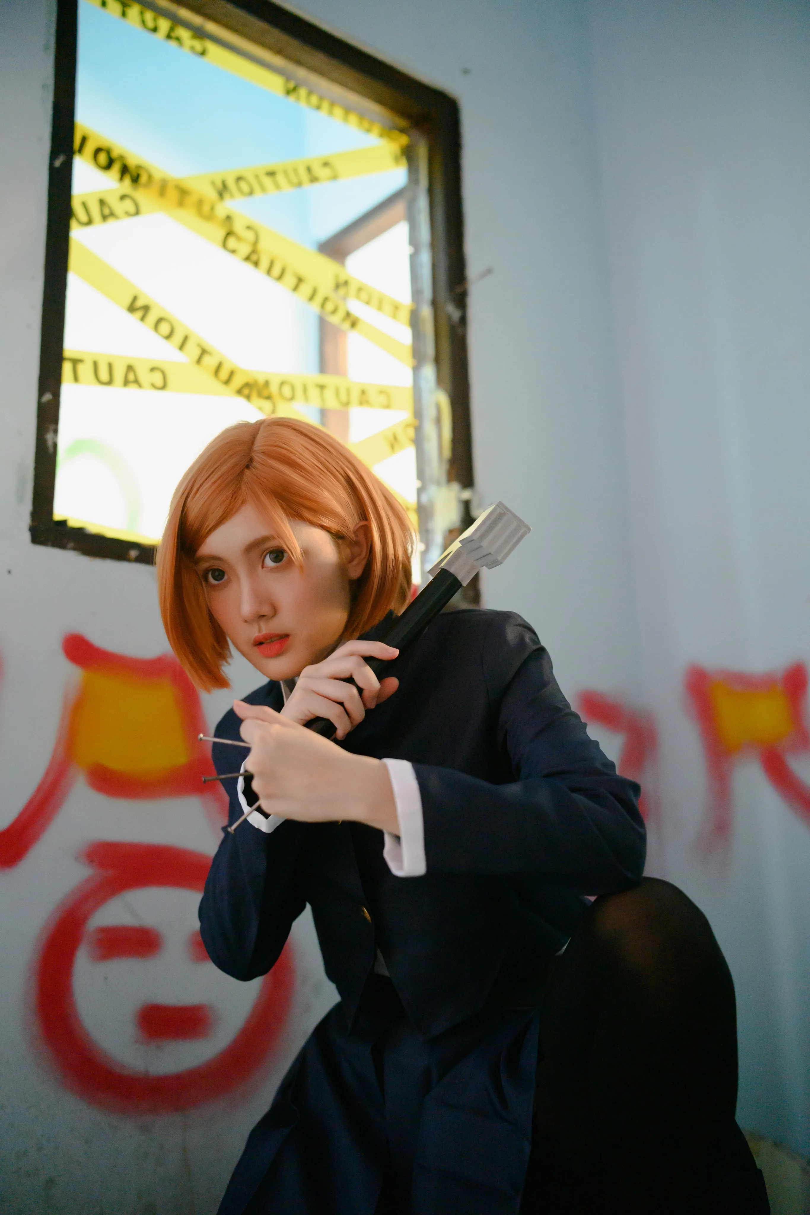 A cosplayer in an orange bob wig crouching with a hammer beneath a window taped with caution tape