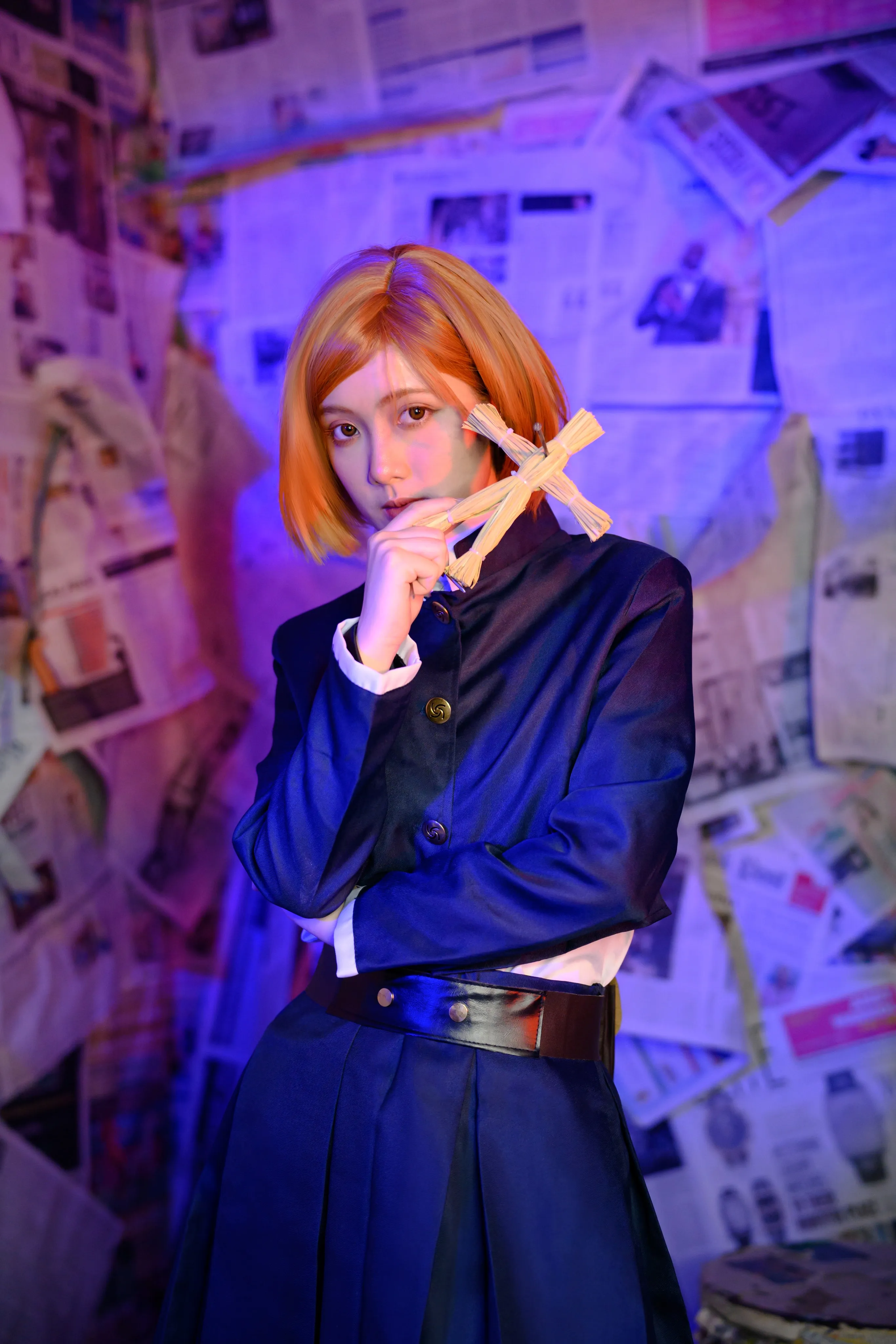 A cosplayer in an orange bob wig holding a straw doll against newspaper-covered walls