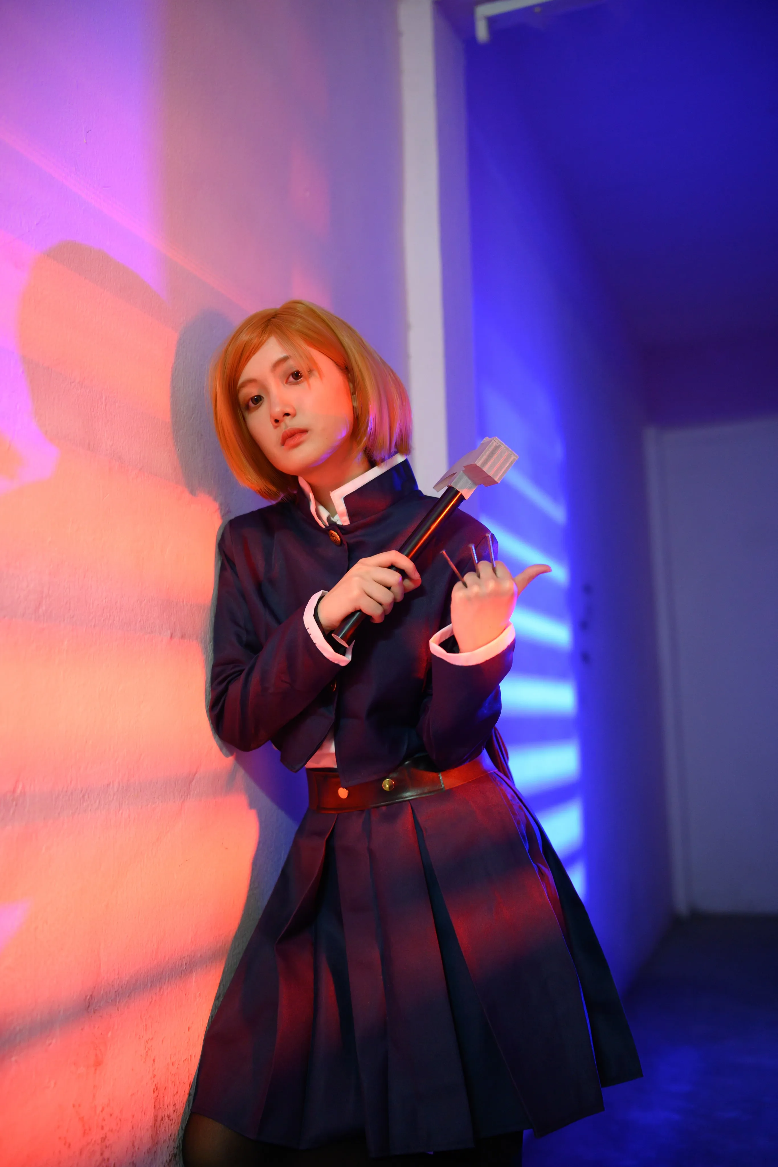 A cosplayer in an orange bob wig holding a hammer and nails against a wall in pink and blue light