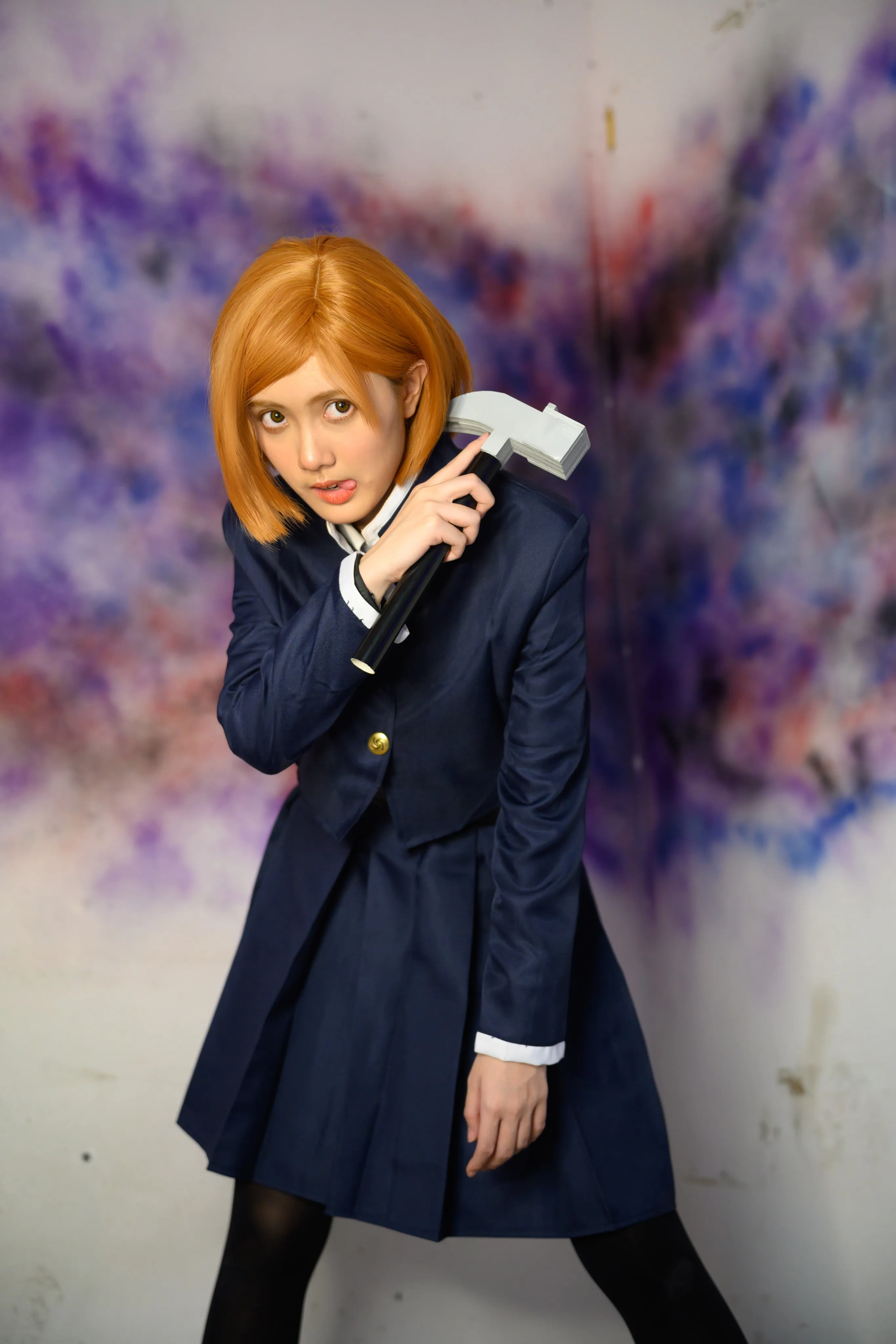 A cosplayer in an orange bob wig holding a hammer against a spray-painted wall