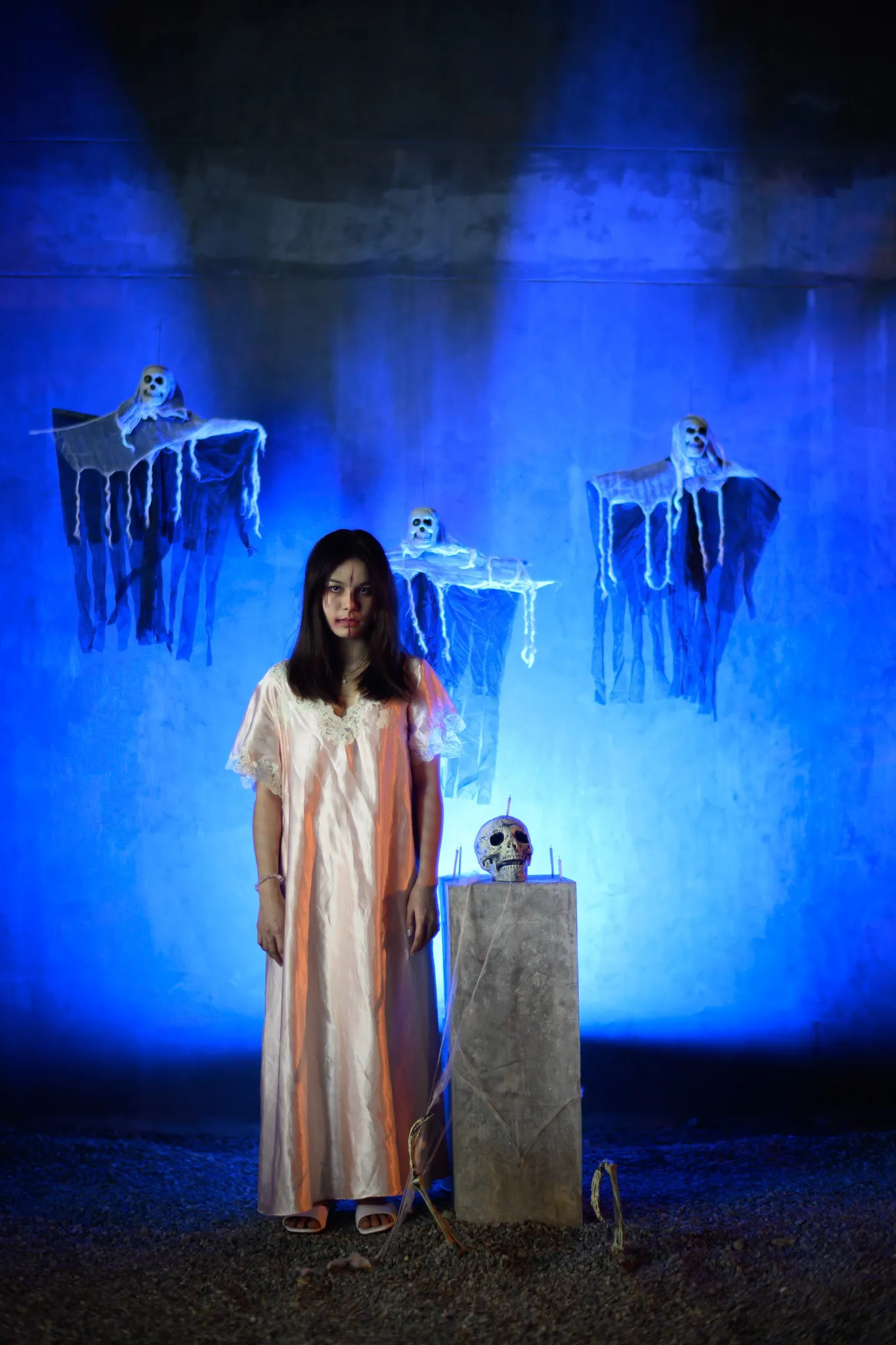 A woman in a pale nightgown standing among hanging skeleton decorations under blue light