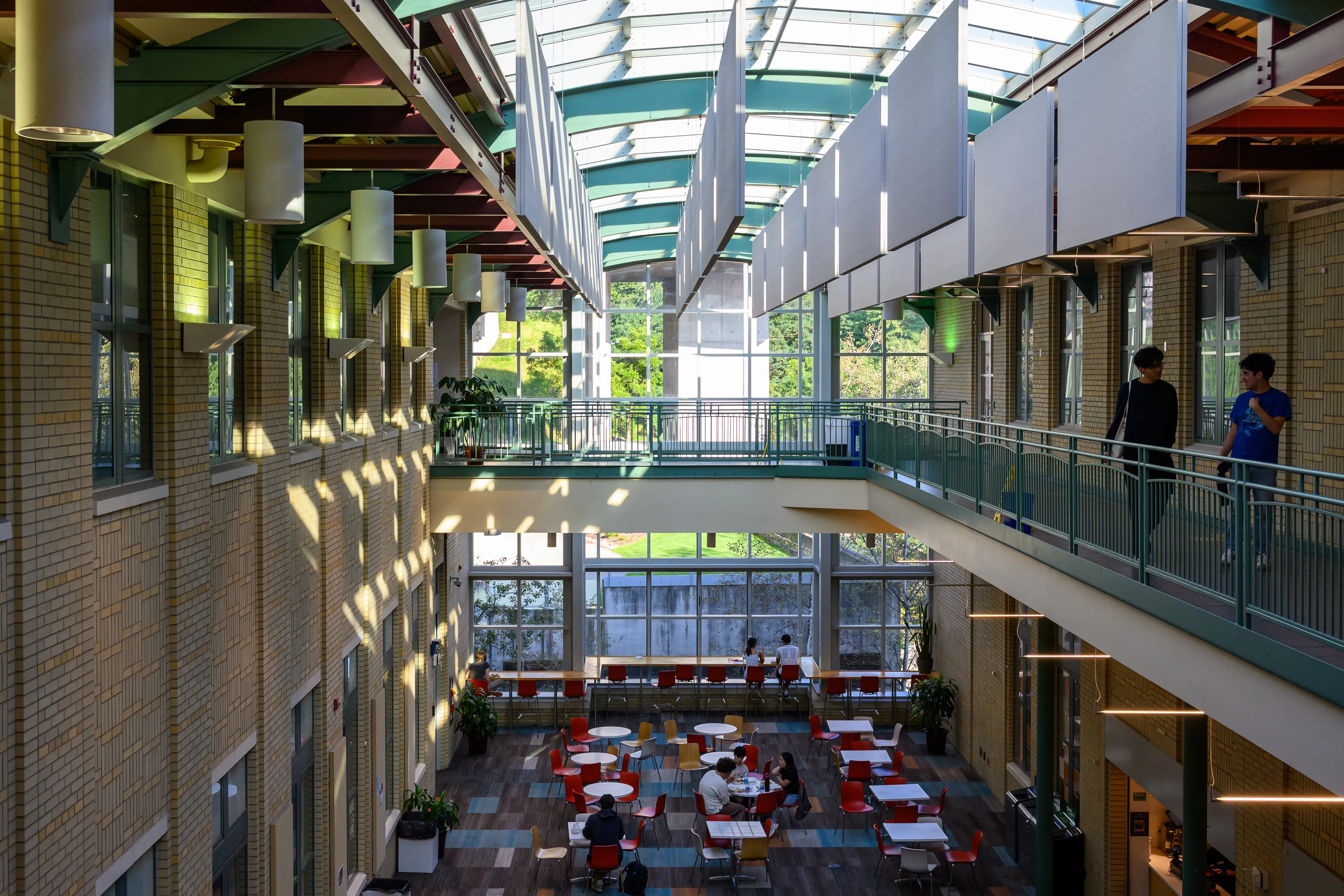 A sunlit multi-story brick atrium under a glass canopy roof hung with white panels, with students crossing a walkway and sitting at red chairs and tables below.
