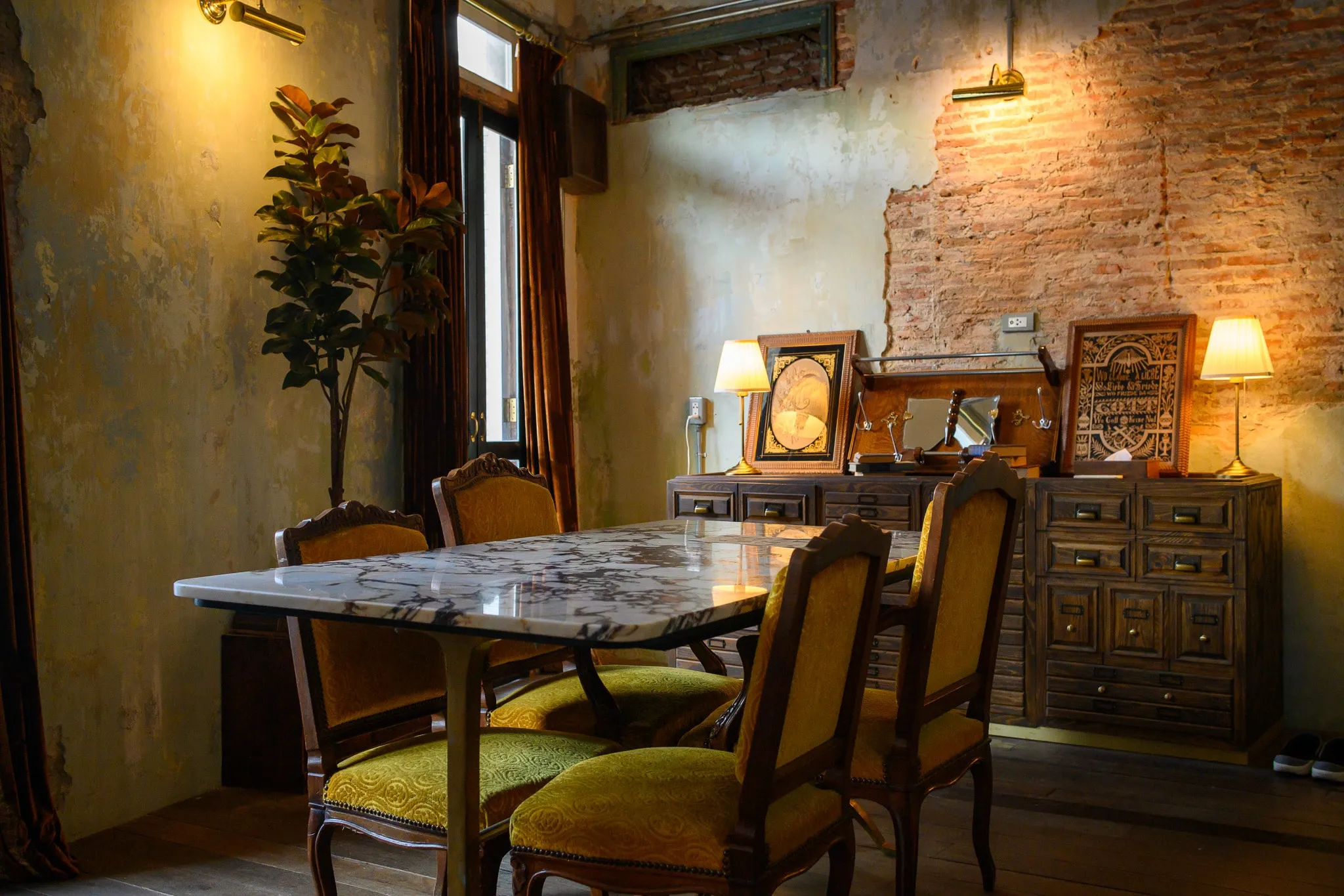 A marble dining table with velvet chairs beside an exposed-brick wall and table lamps