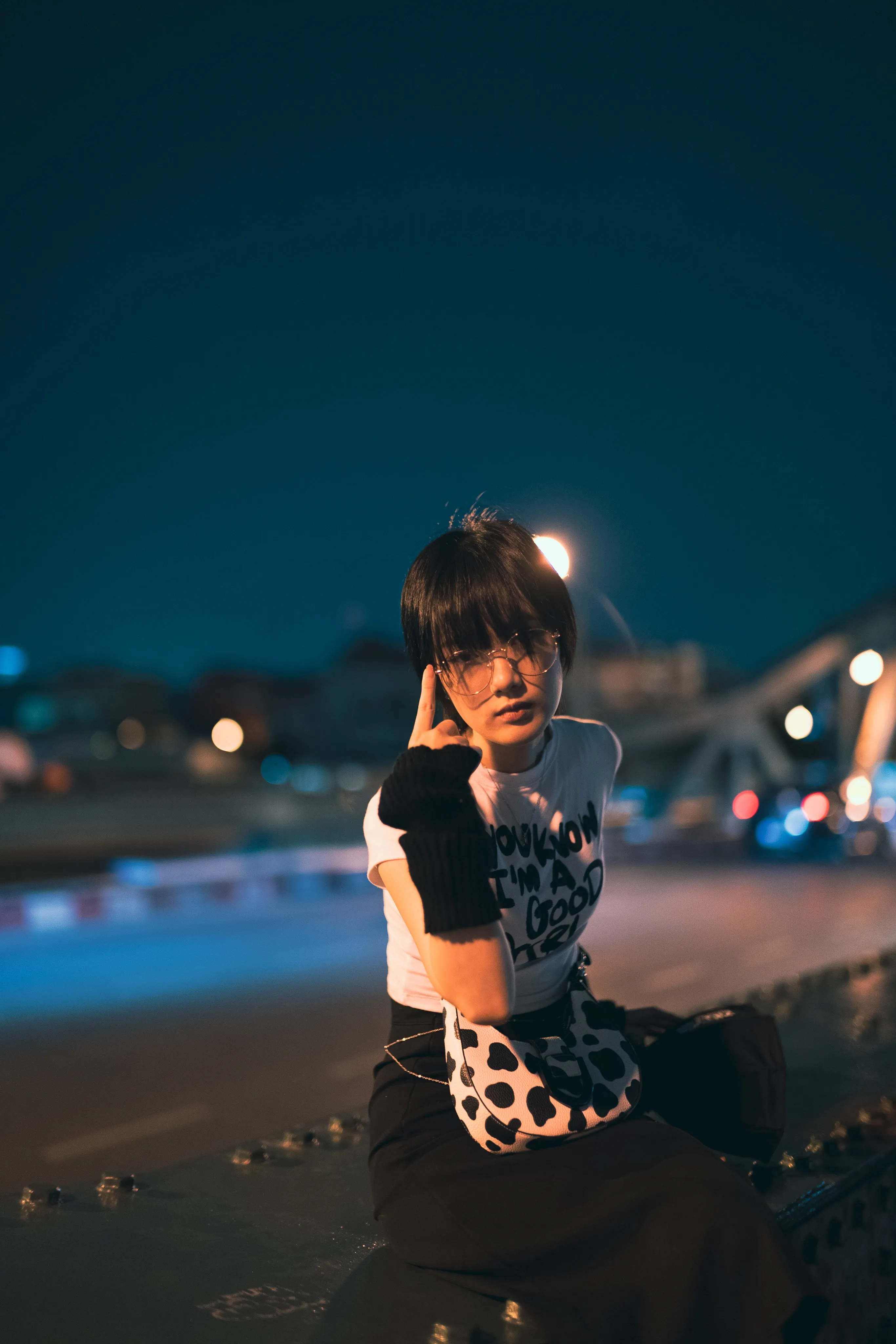 A woman in a graphic T-shirt and black gloves seated on a steel bridge at night, city lights blurred behind her