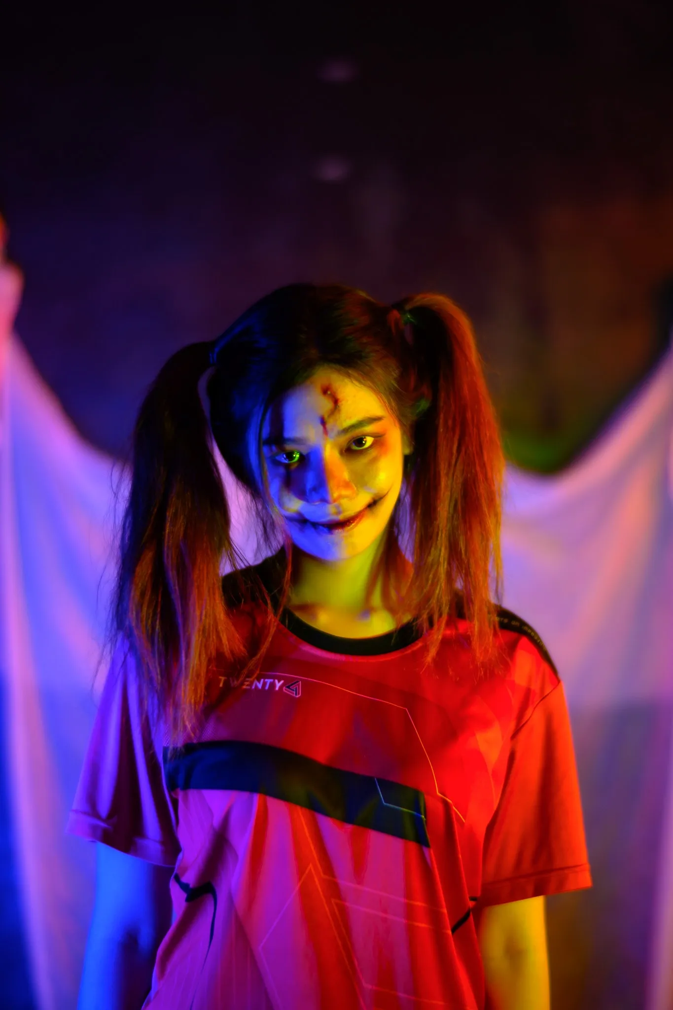 A person in pigtails and a red jersey with painted horror makeup smiling under colored light