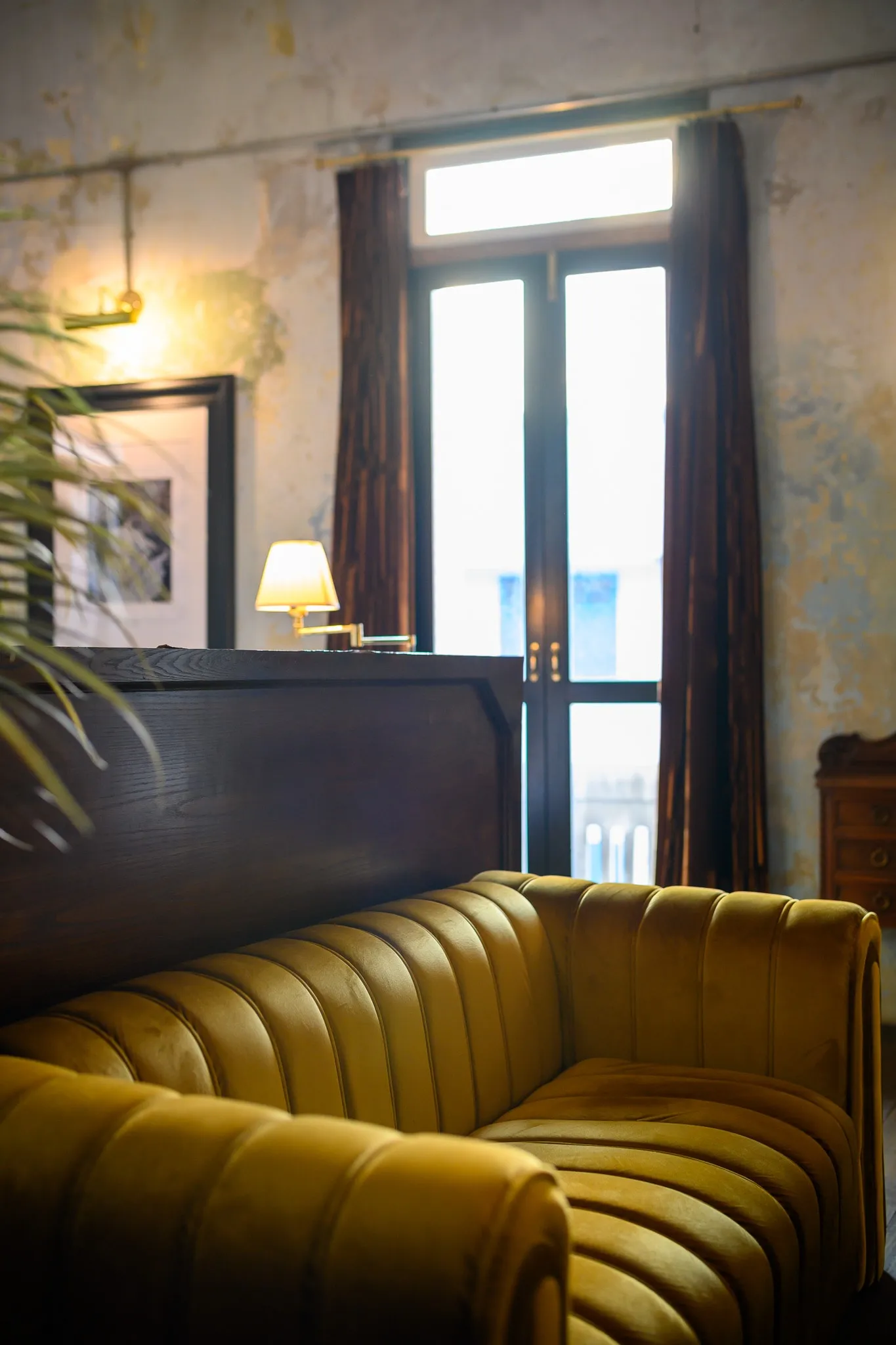 A curved mustard velvet sofa beneath tall French windows and a wall lamp