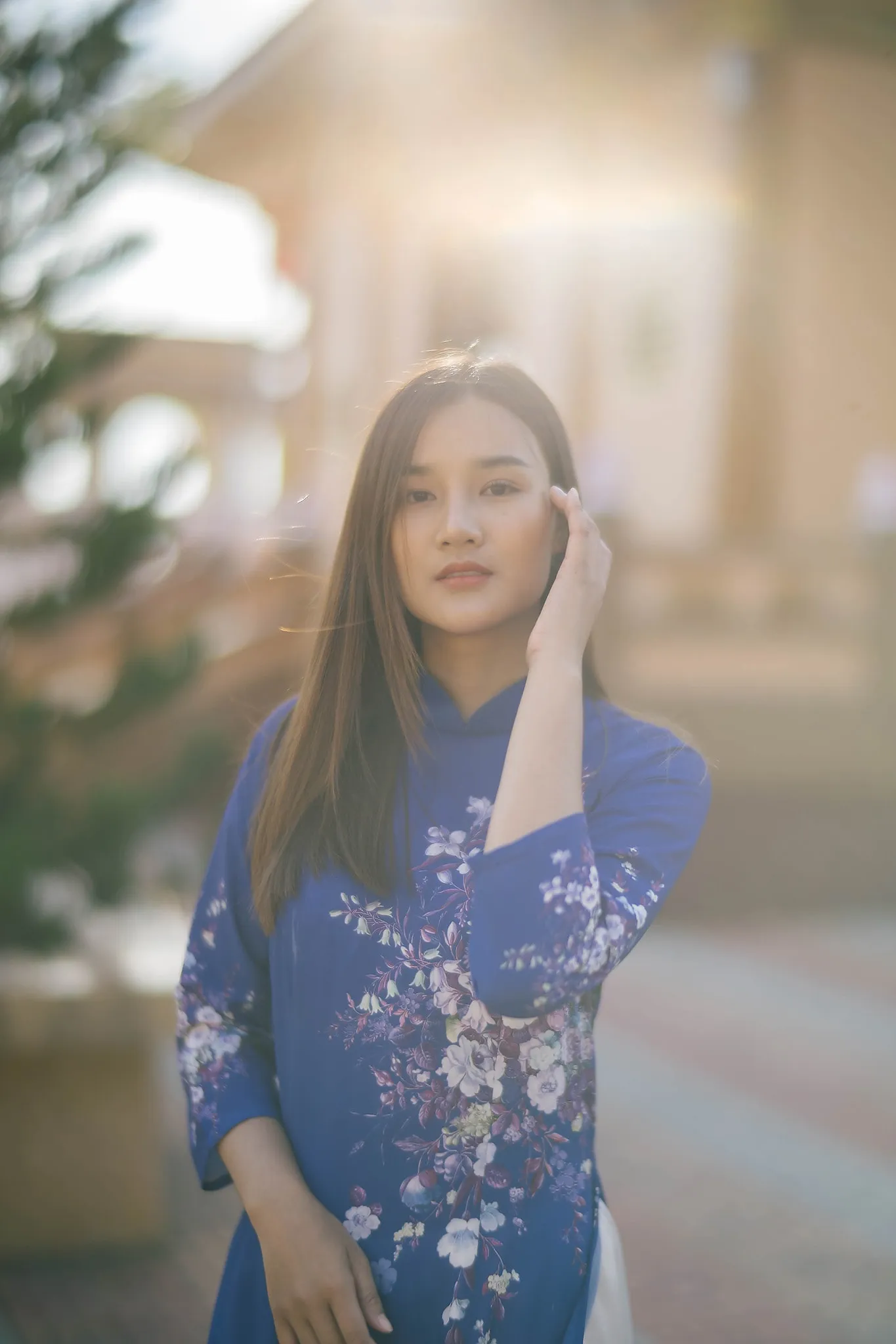 A woman in a blue floral ao dai touching her hair in warm backlight