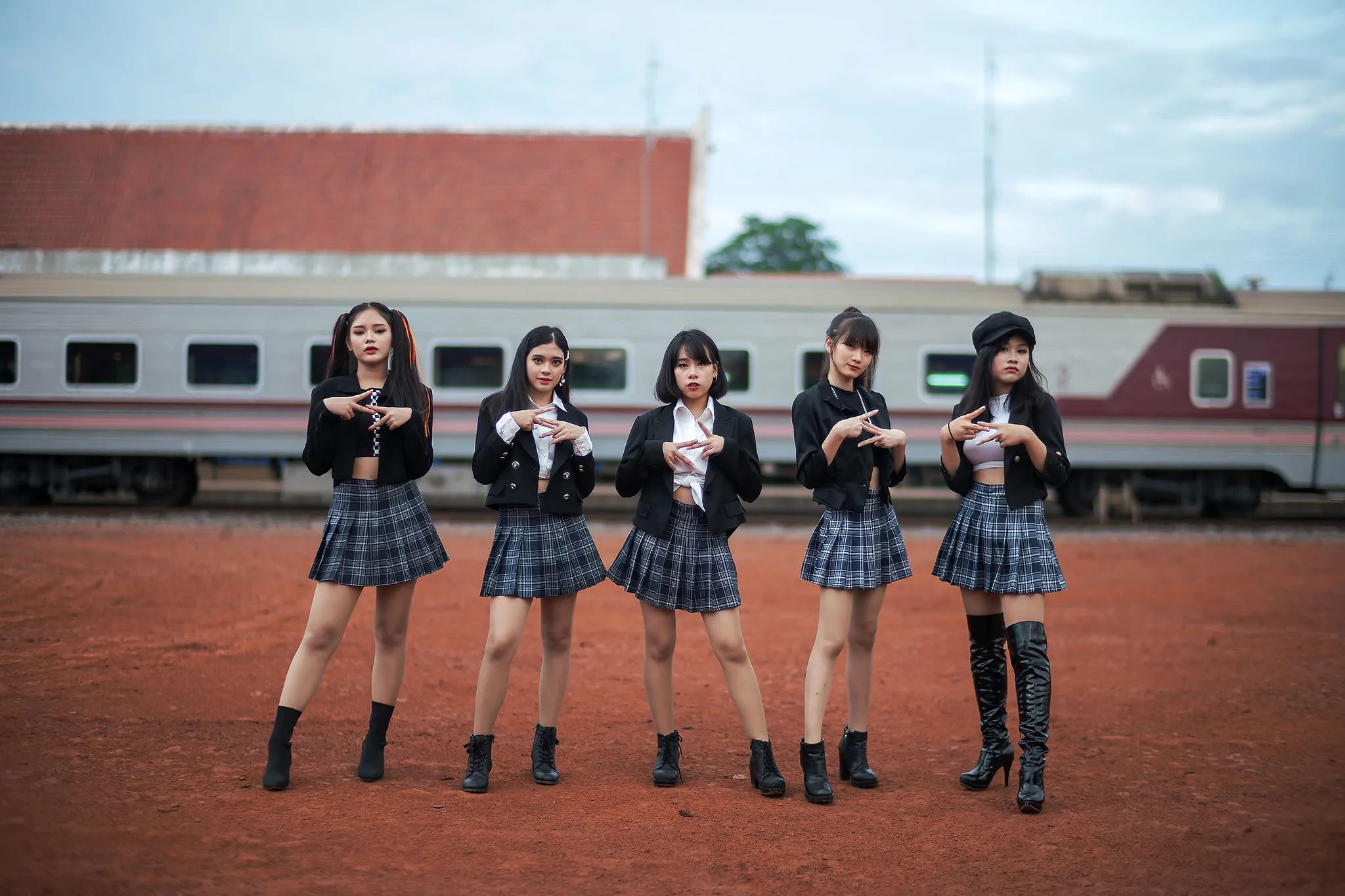 Five performers in black jackets and plaid skirts posing on red earth in front of a passenger train