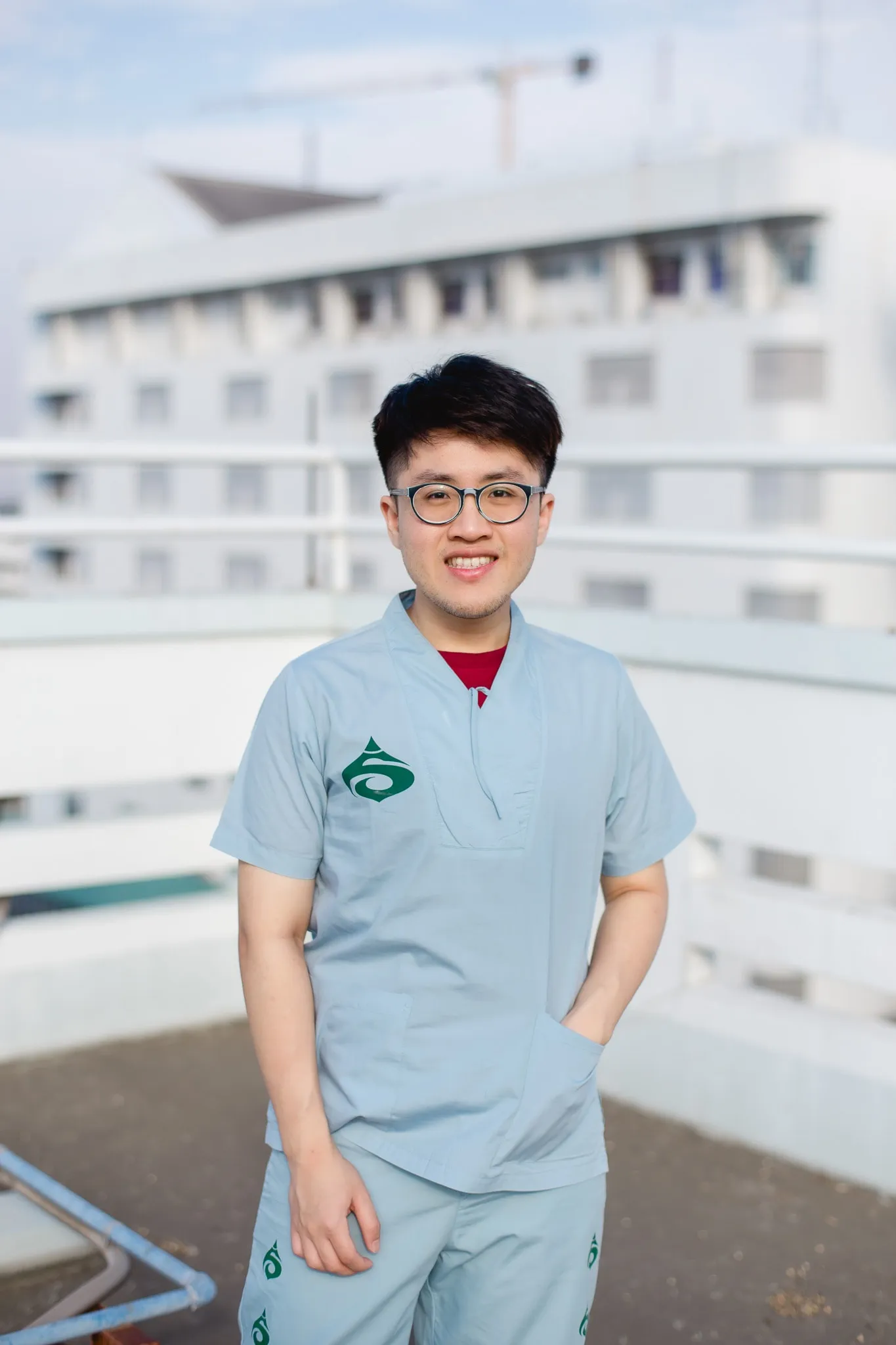A smiling person with glasses in light blue medical scrubs standing on a rooftop