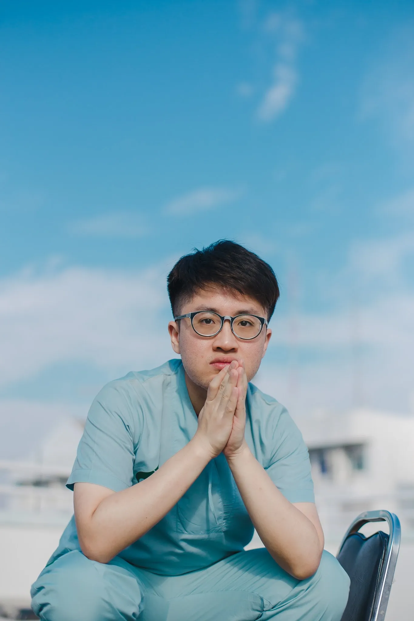 A person with glasses in medical scrubs sitting with hands pressed together beneath a bright blue sky