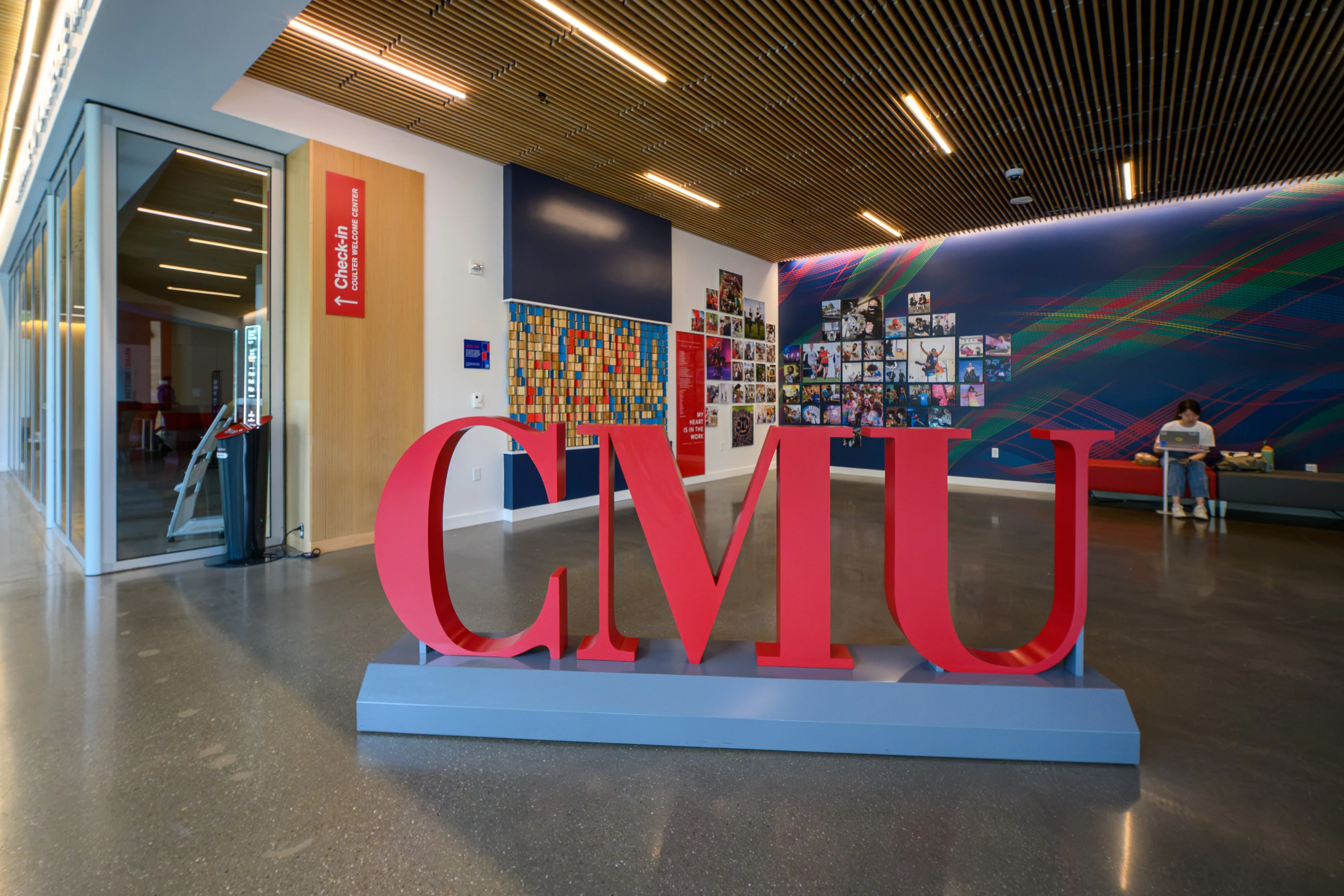 A red freestanding "CMU" sign in a bright, art-filled campus lobby.