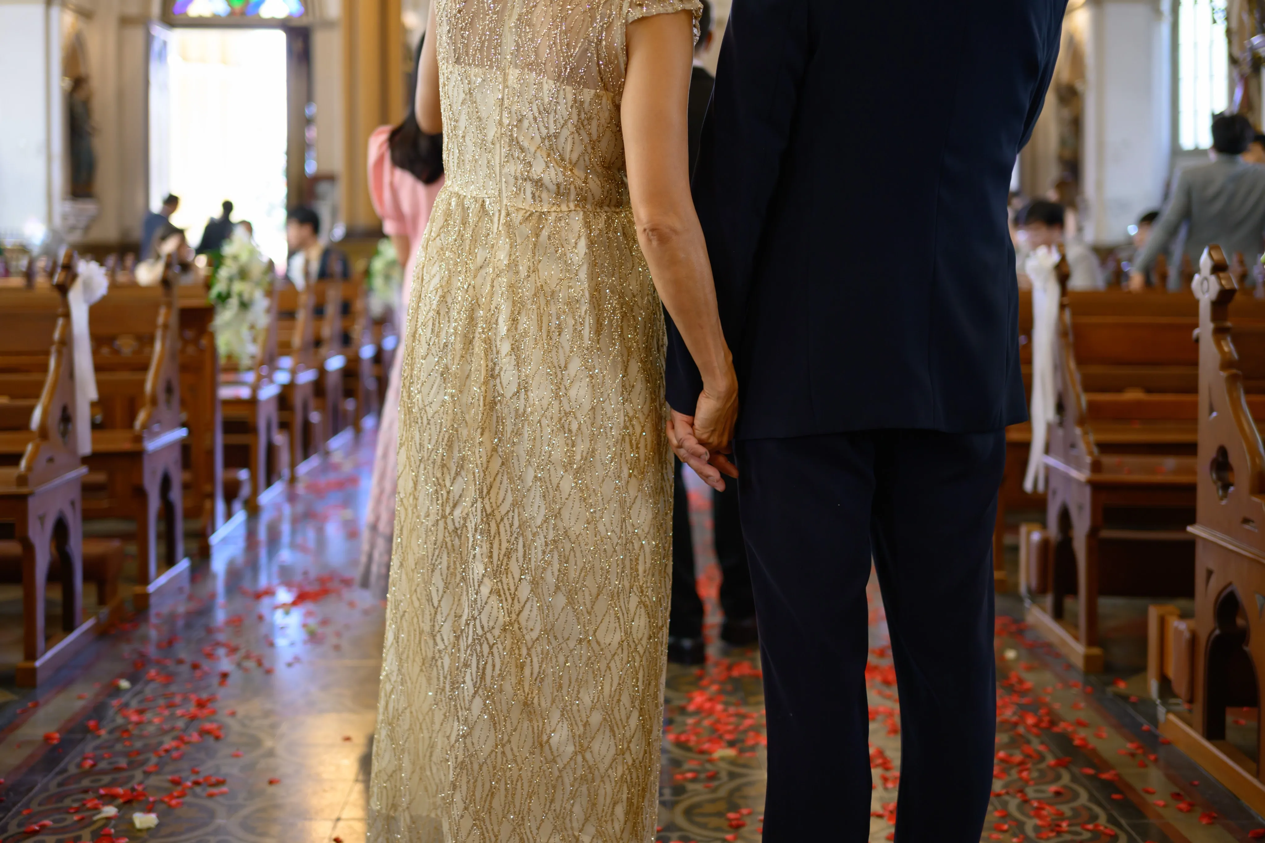 A couple in a gold sequined gown and a dark suit holding hands in a church aisle scattered with red rose petals
