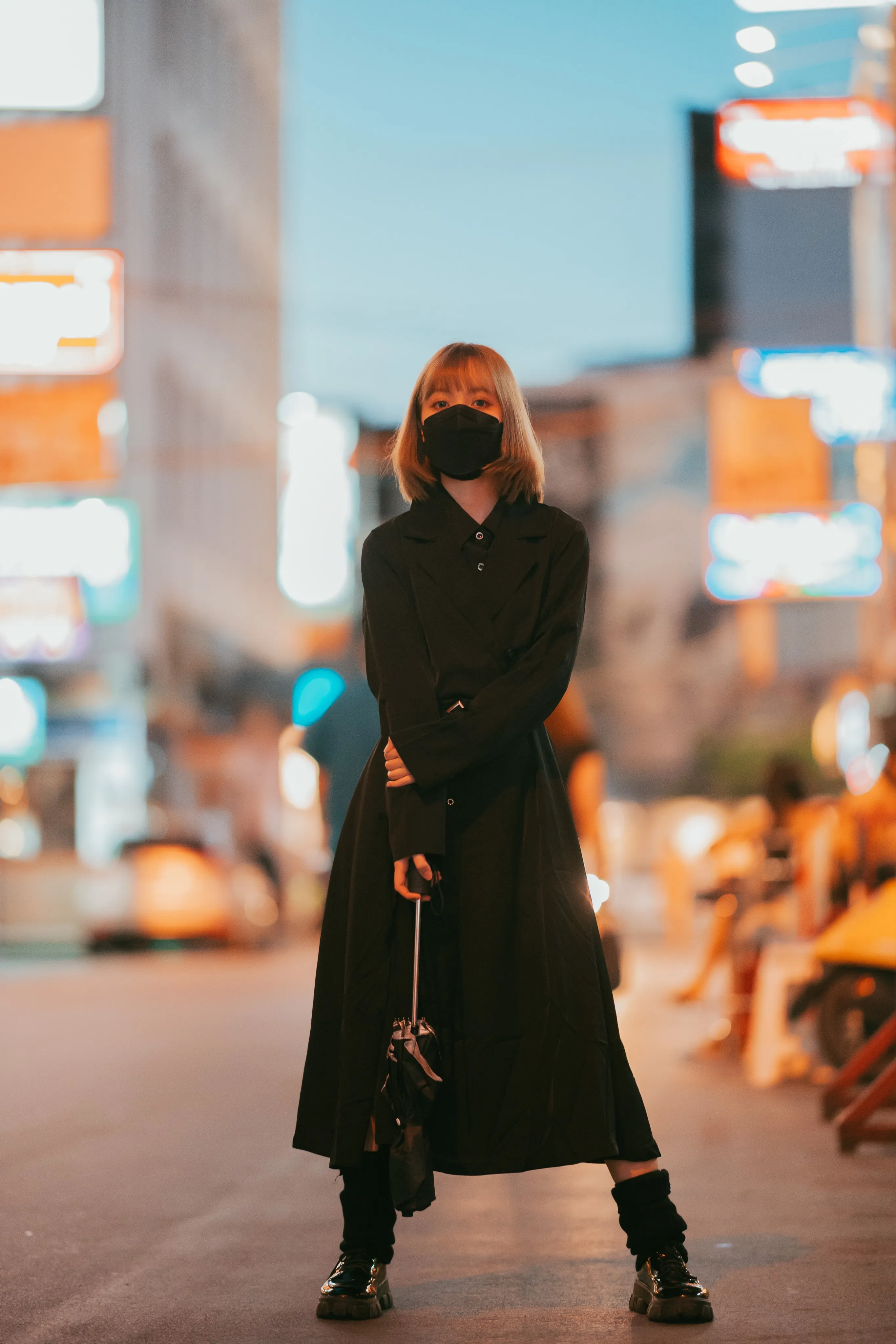 A person in a black mask and long black coat dress holding a folded umbrella on a street lined with glowing neon signs at dusk