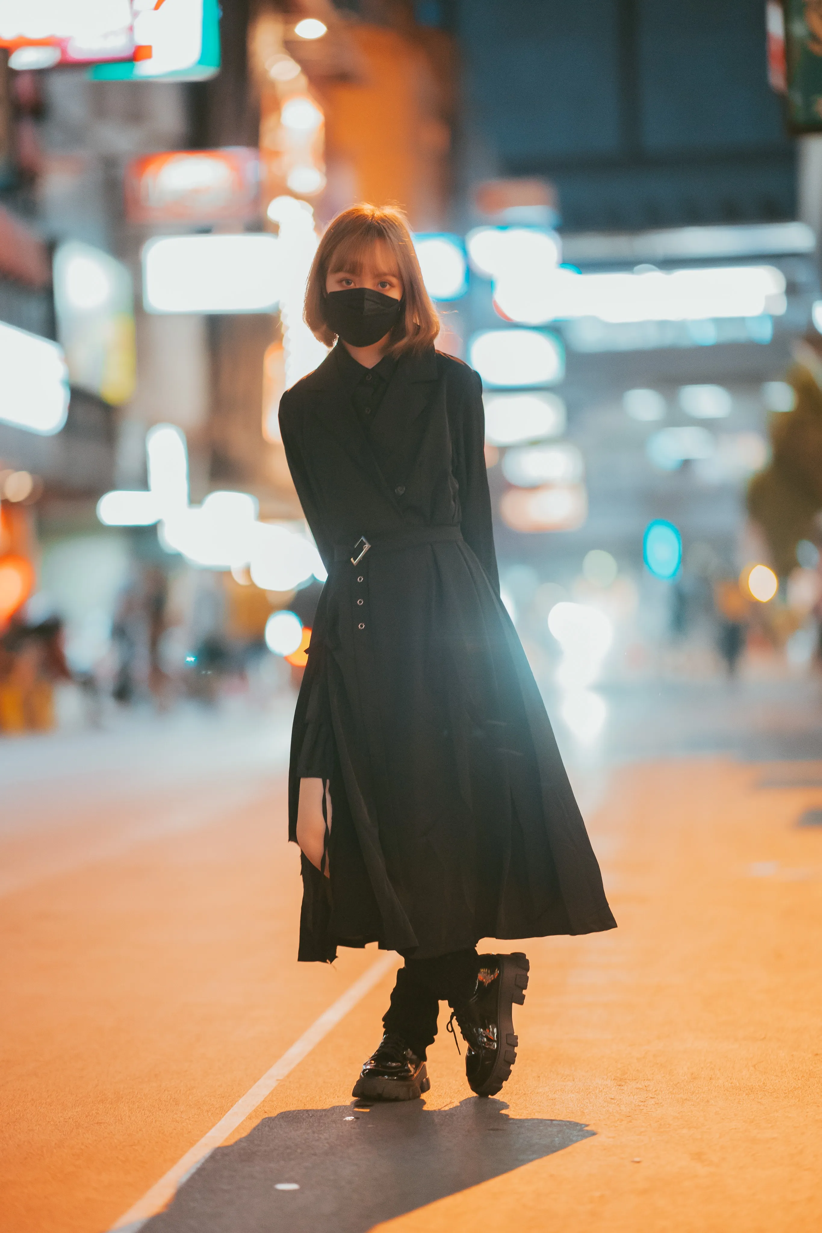A person in a black mask and belted black coat dress walking along an amber-lit street at night beneath blurred neon signs
