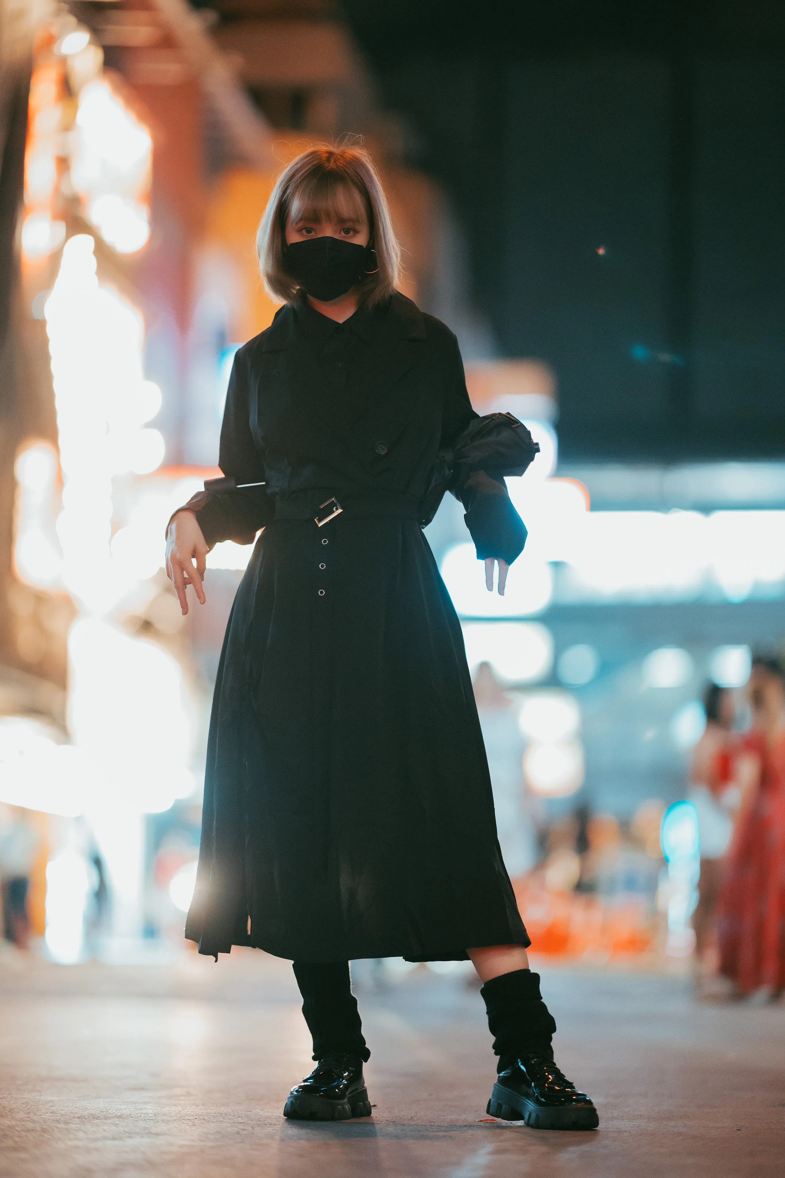 A person in a black mask and black coat dress posing with arms raised on a neon-lit street at night, a folded umbrella tucked at one elbow