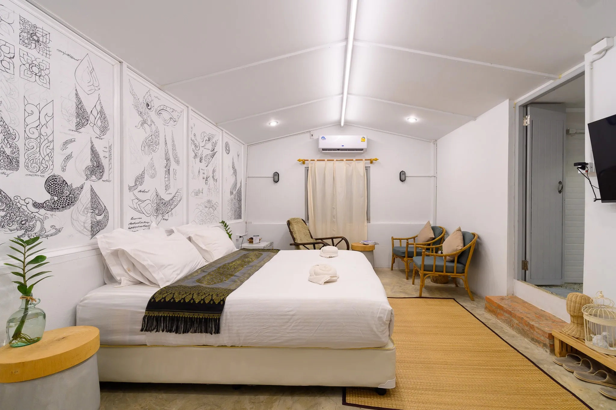 A bright hotel room with line-drawn murals, a white bed, and rattan armchairs