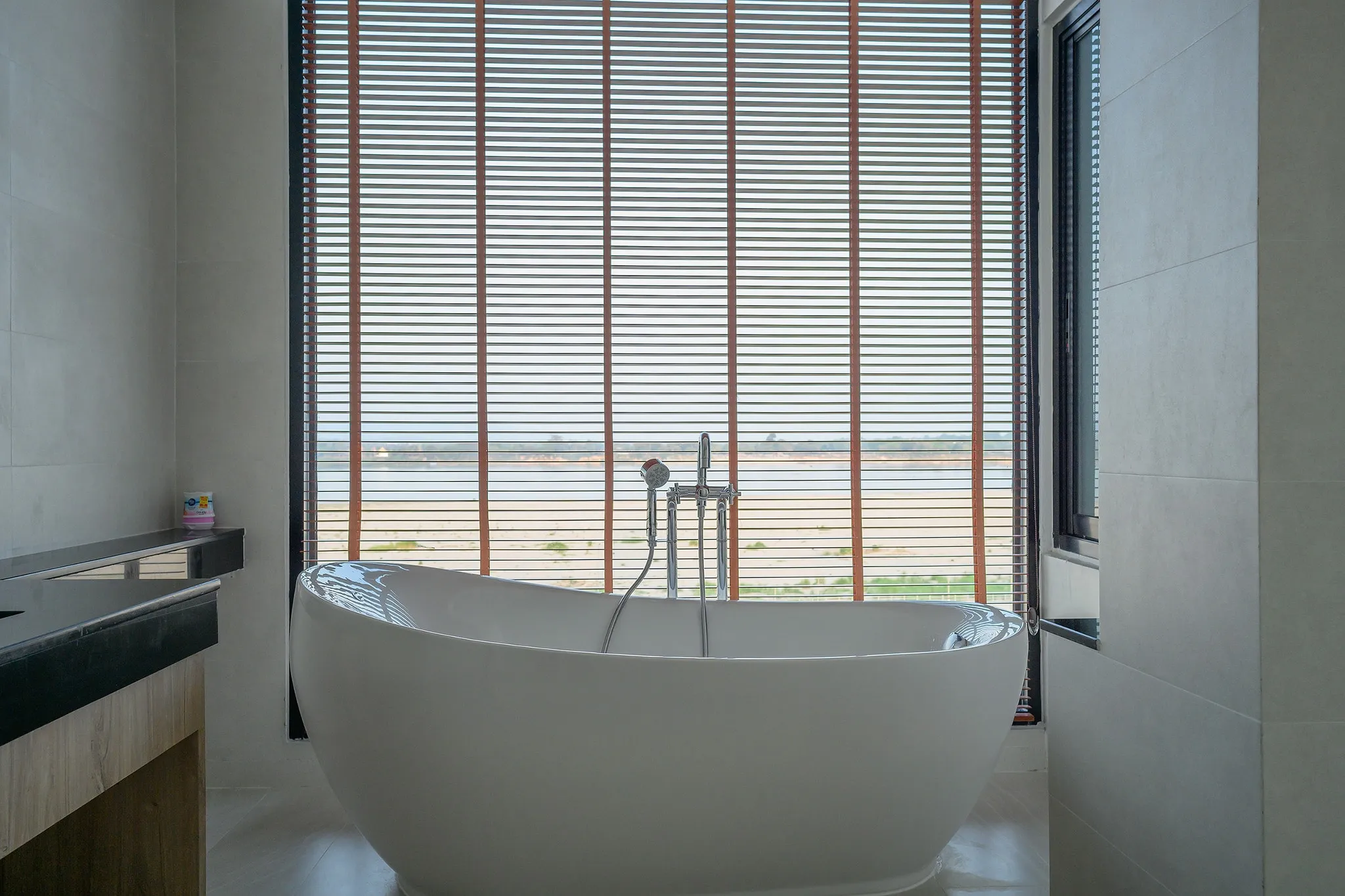 A freestanding white bathtub in front of a slatted blind overlooking a river