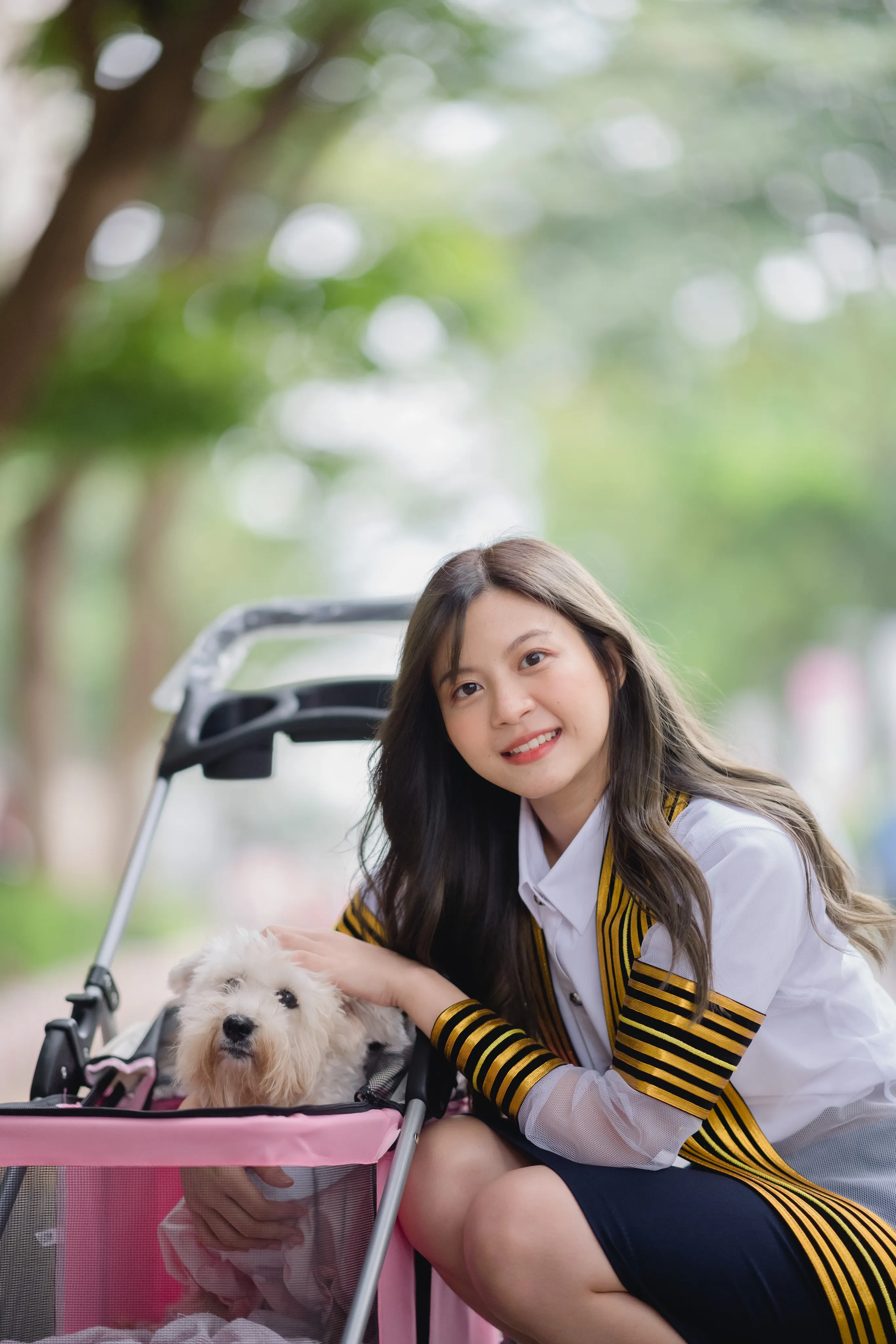 A smiling graduate in academic dress crouching beside a white dog in a pink pet stroller