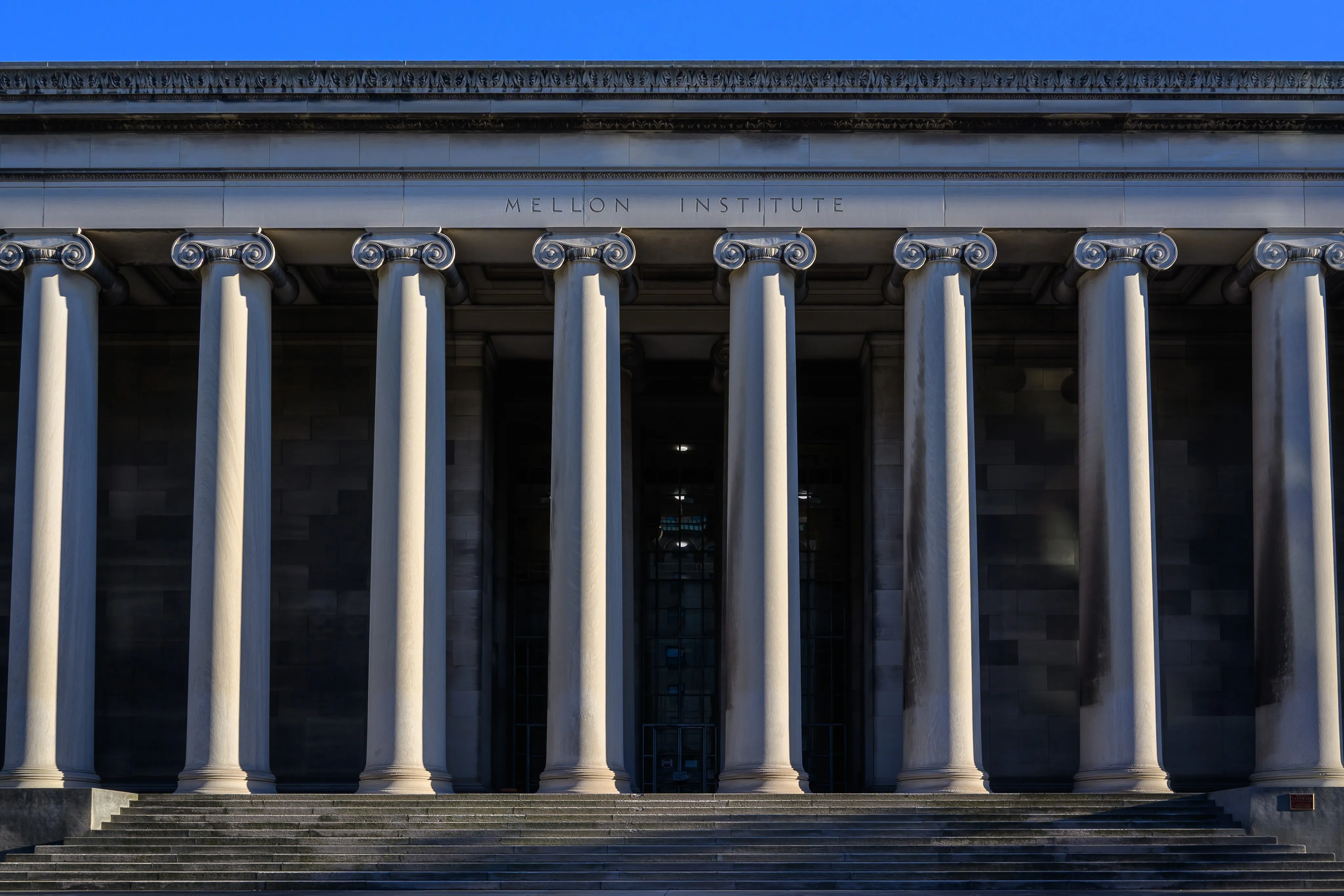 The Ionic columns and broad stone steps of the Mellon Institute facade, its name carved in the entablature, under a deep blue sky.