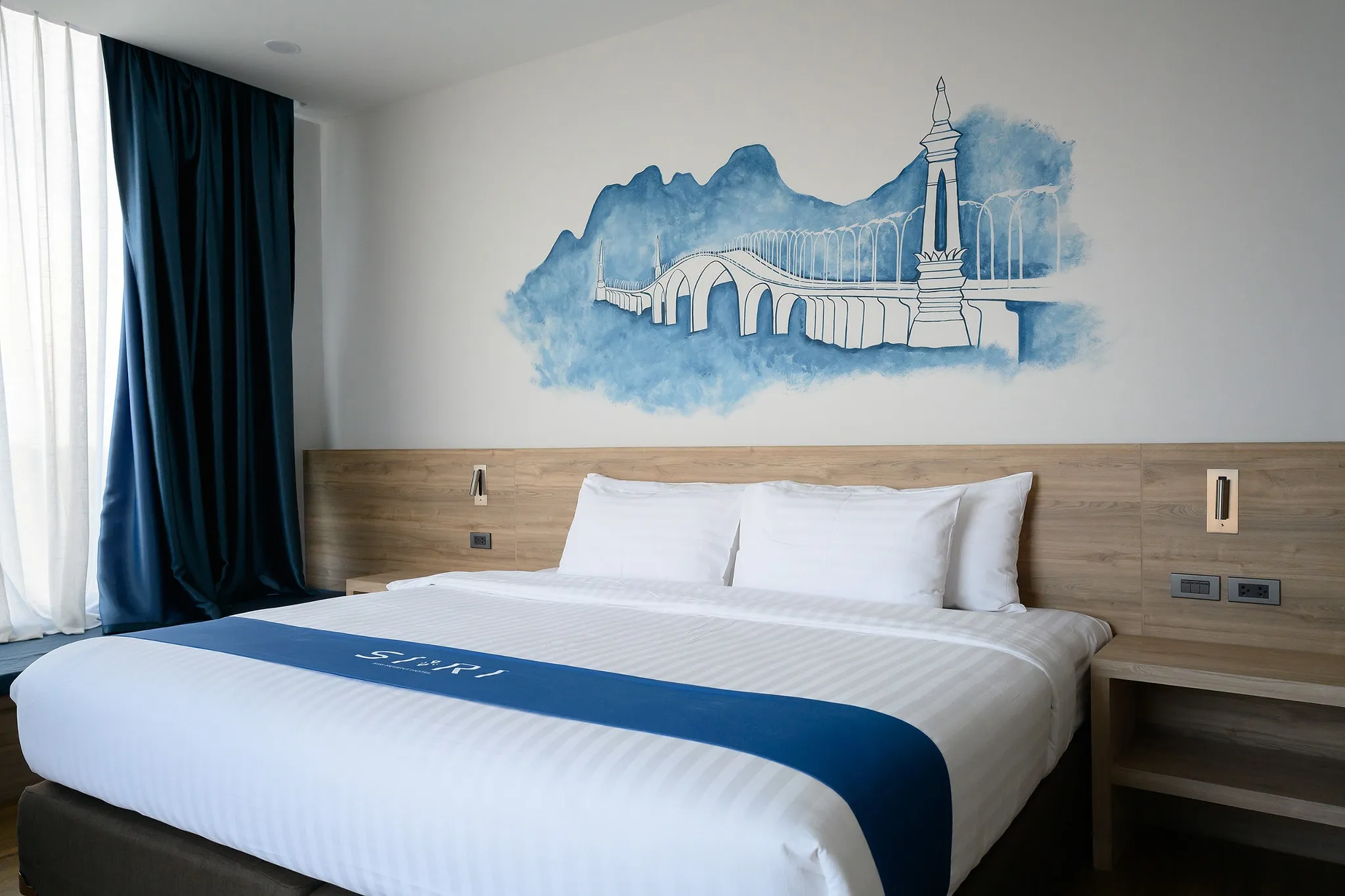 A hotel bedroom with a blue mural of a bridge and landmark tower above the bed