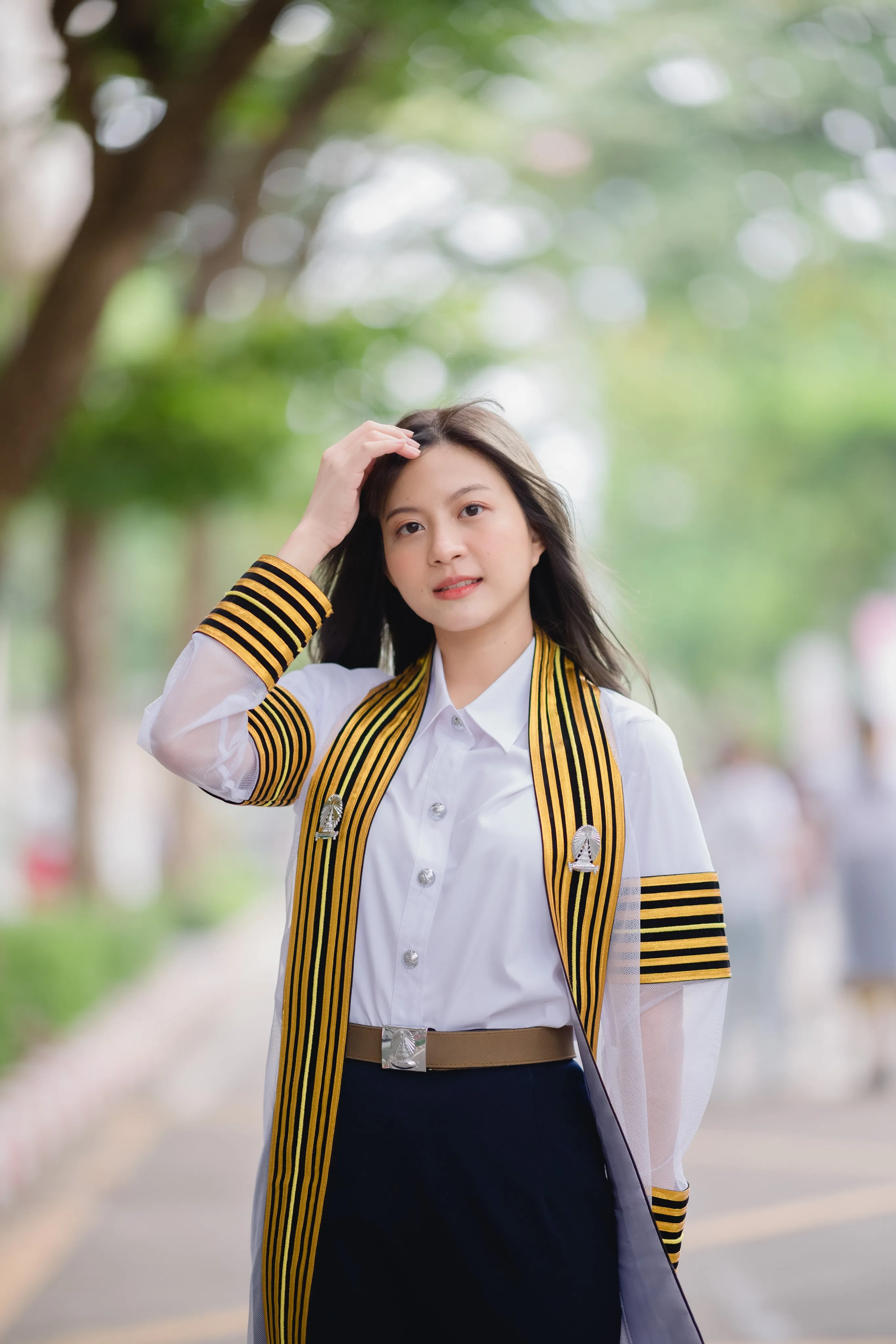 A graduate in a striped black-and-gold academic gown touching her hair on a tree-lined campus road