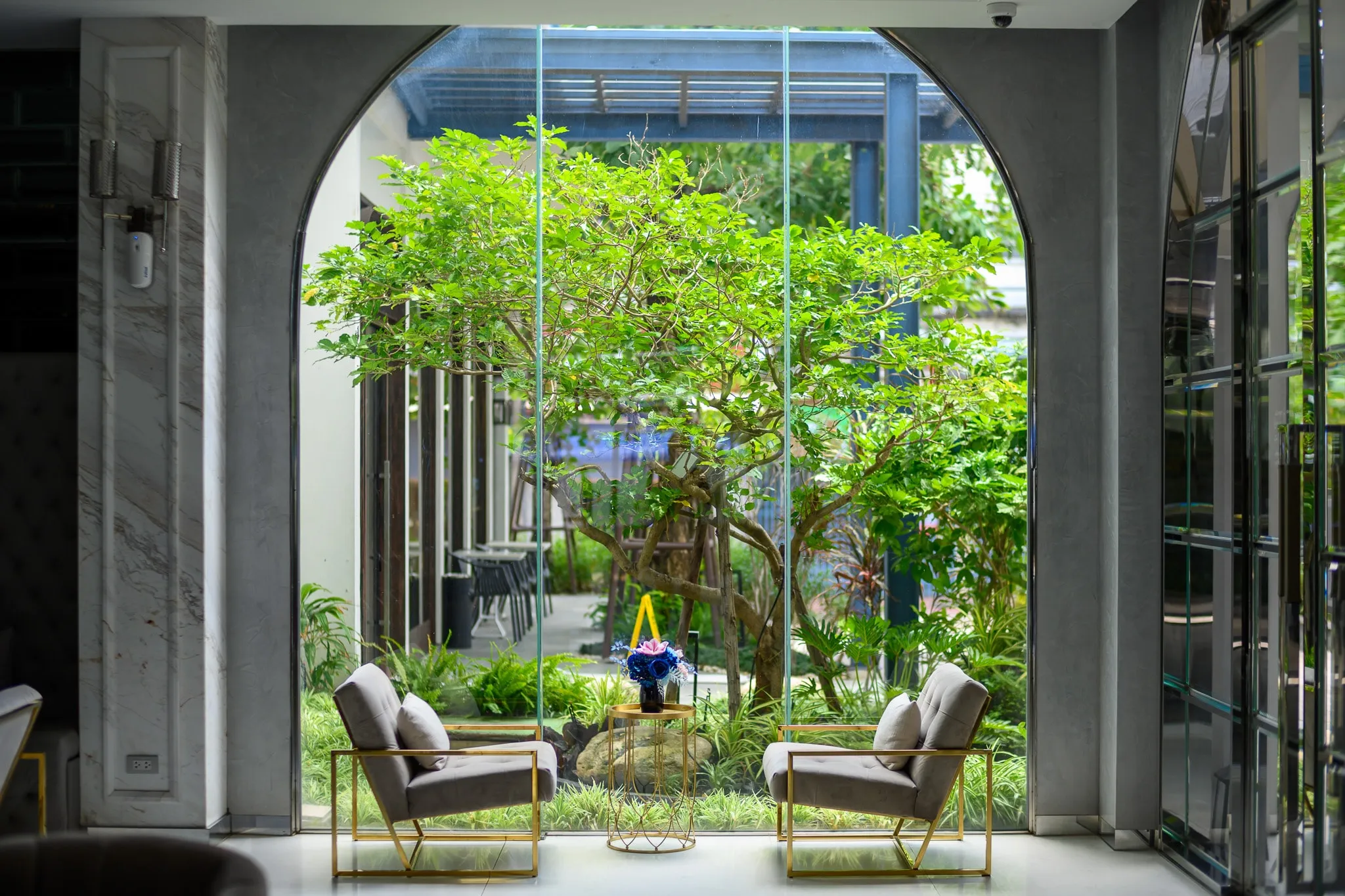 Two lounge chairs facing a lush inner garden through a tall arched glass window