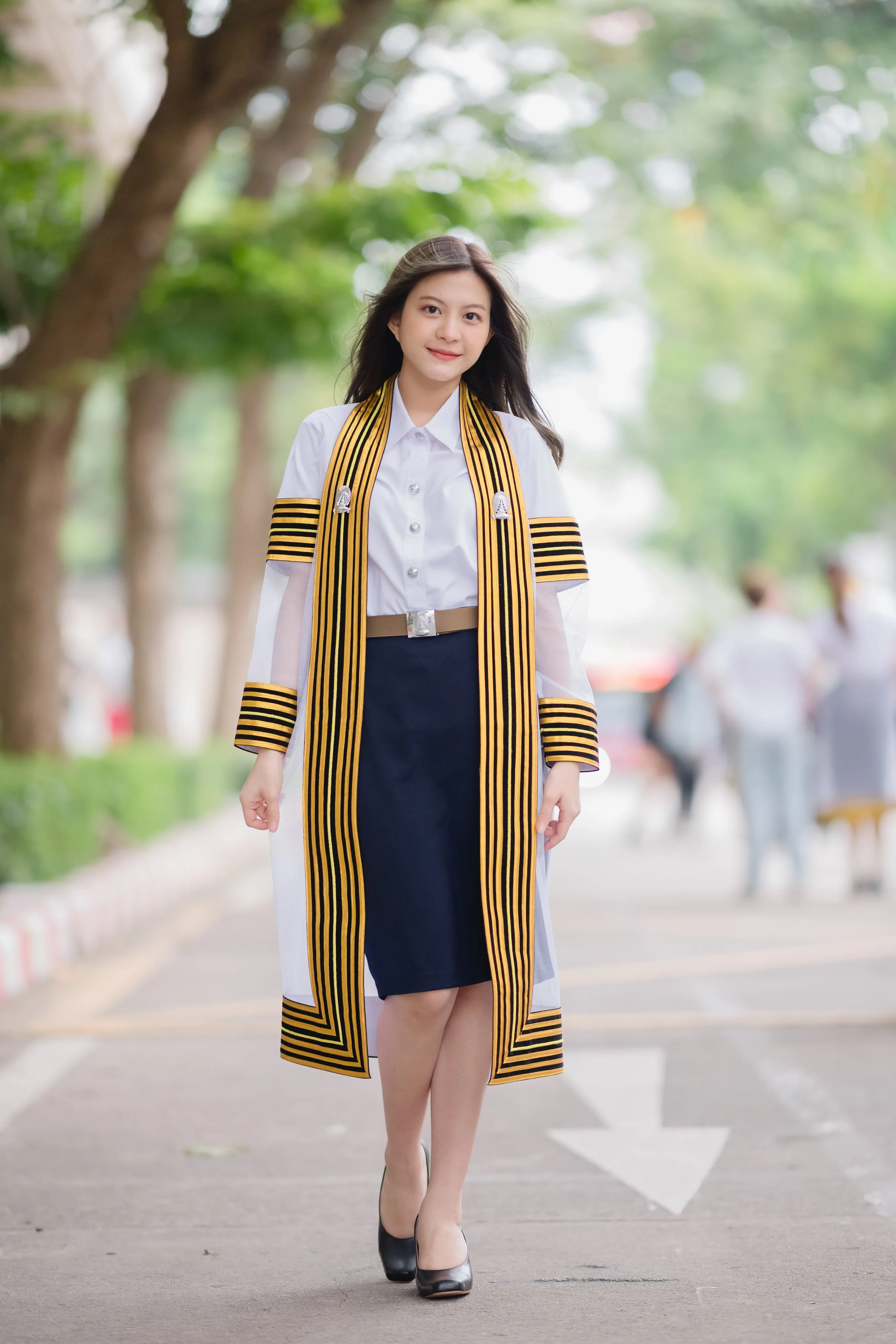 A graduate in a striped black-and-gold academic gown walking toward the camera along a tree-lined road