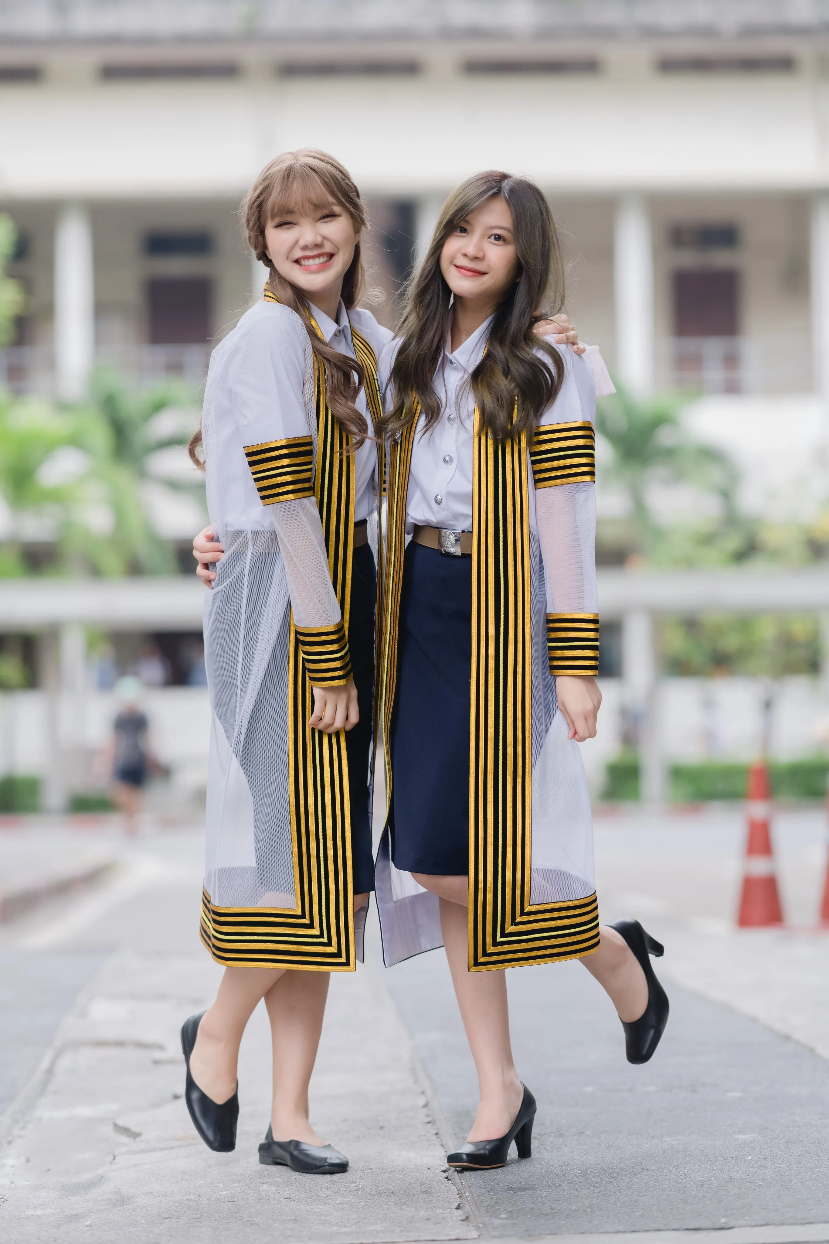 Two smiling graduates in striped academic gowns posing arm in arm in front of a campus building