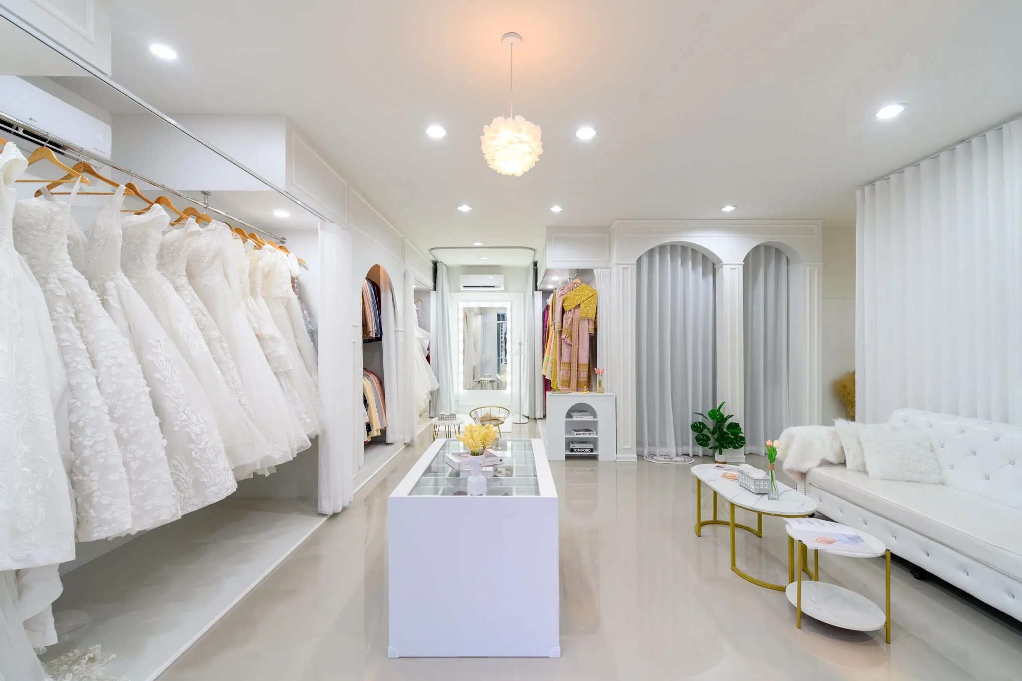 A white bridal studio with racks of wedding gowns, a glass display counter, and a sofa